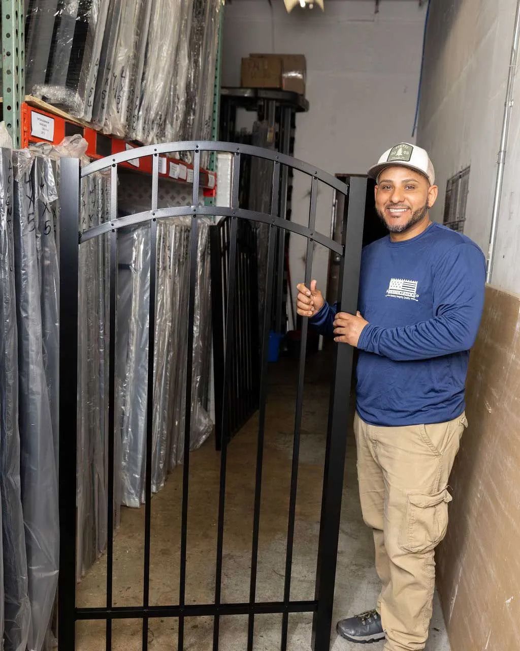 Man in blue shirt and tan pants smiles, holding black metal gate in warehouse setting.