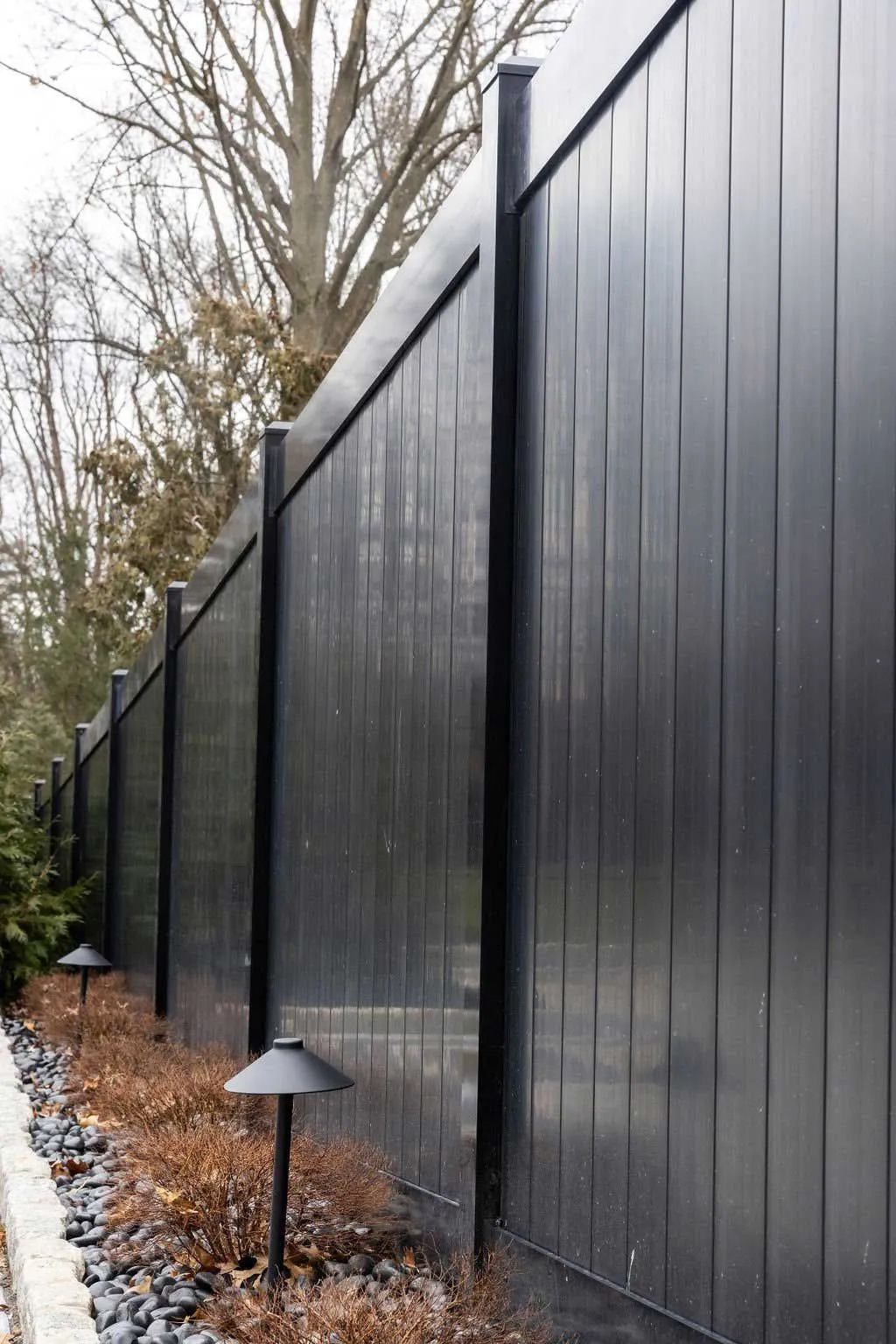 Black vertical-slat fence with black posts. Ground-level landscape lighting illuminates the base along a stone border.