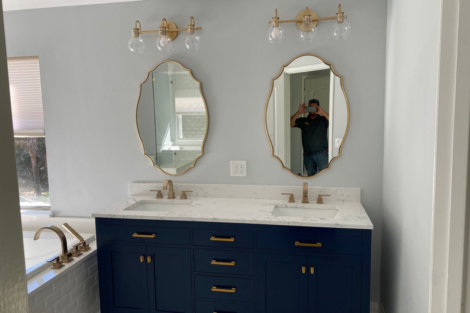 Bathroom with blue vanity, gold fixtures, oval mirrors, and a person taking a photo.
