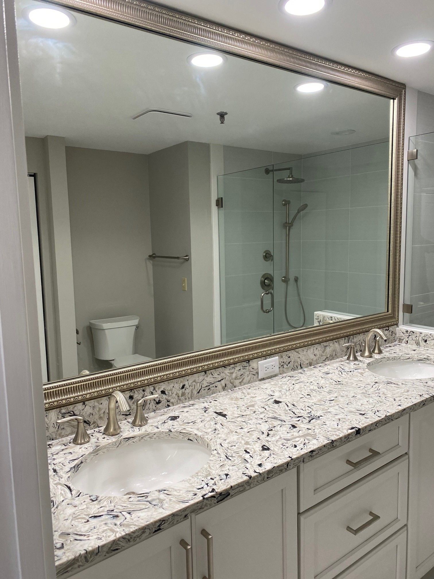 Bathroom with large mirror reflecting a toilet, shower, and double vanity with white cabinets and speckled countertop.