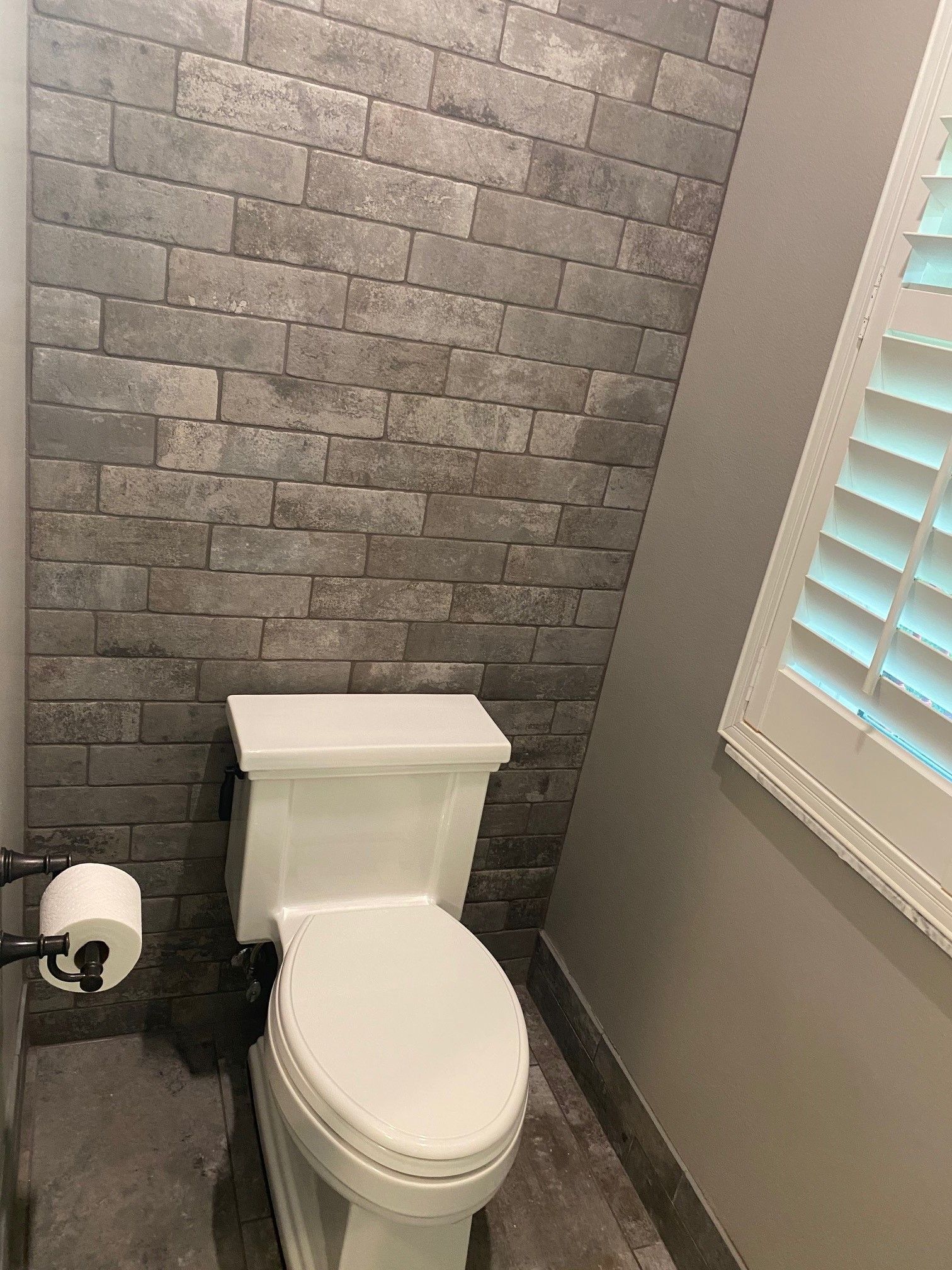 White toilet in a small bathroom with gray brick-style accent wall and a shuttered window.