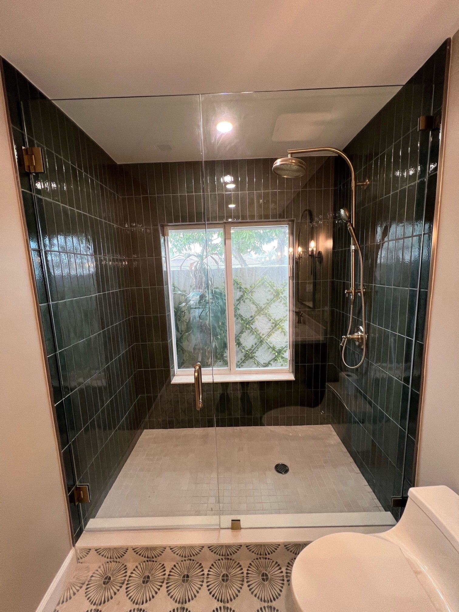 A modern shower with dark green tile walls, a glass door, and a window, with a toilet in front.