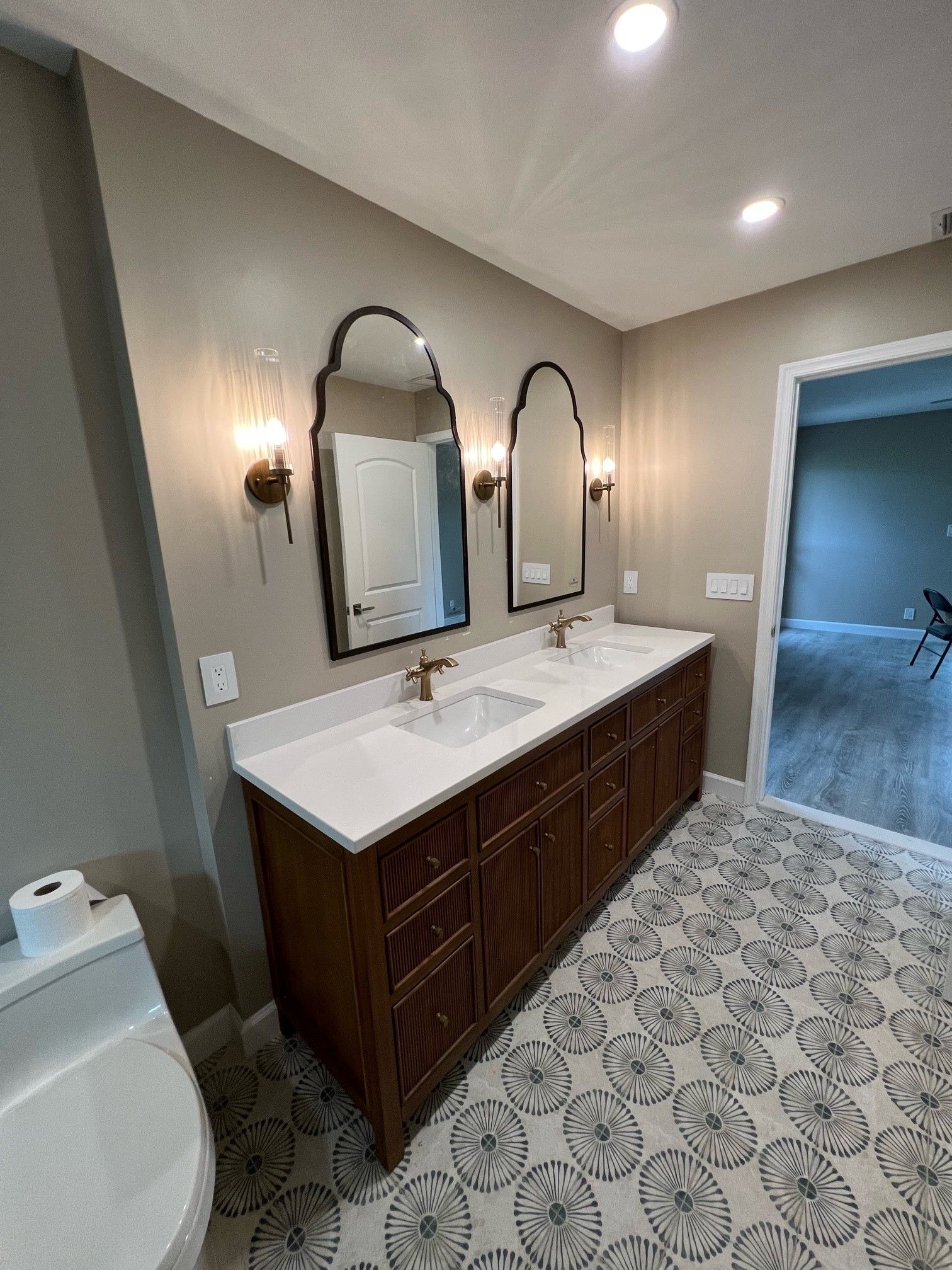 Bathroom with brown vanity, white countertop, patterned floor, arched mirrors, and sconces.