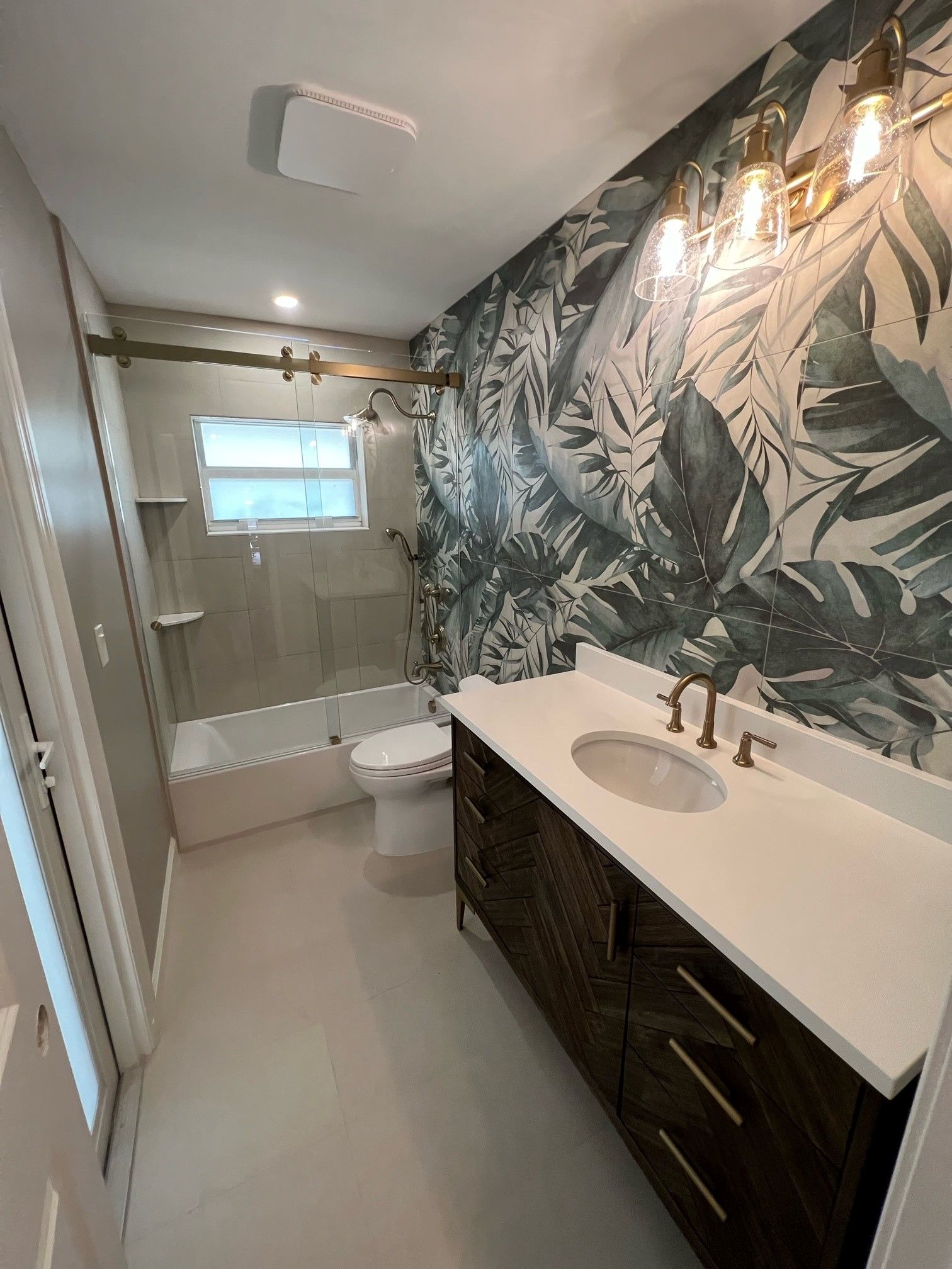 Bathroom with tropical leaf wallpaper, dark vanity, white countertop, and gold fixtures.