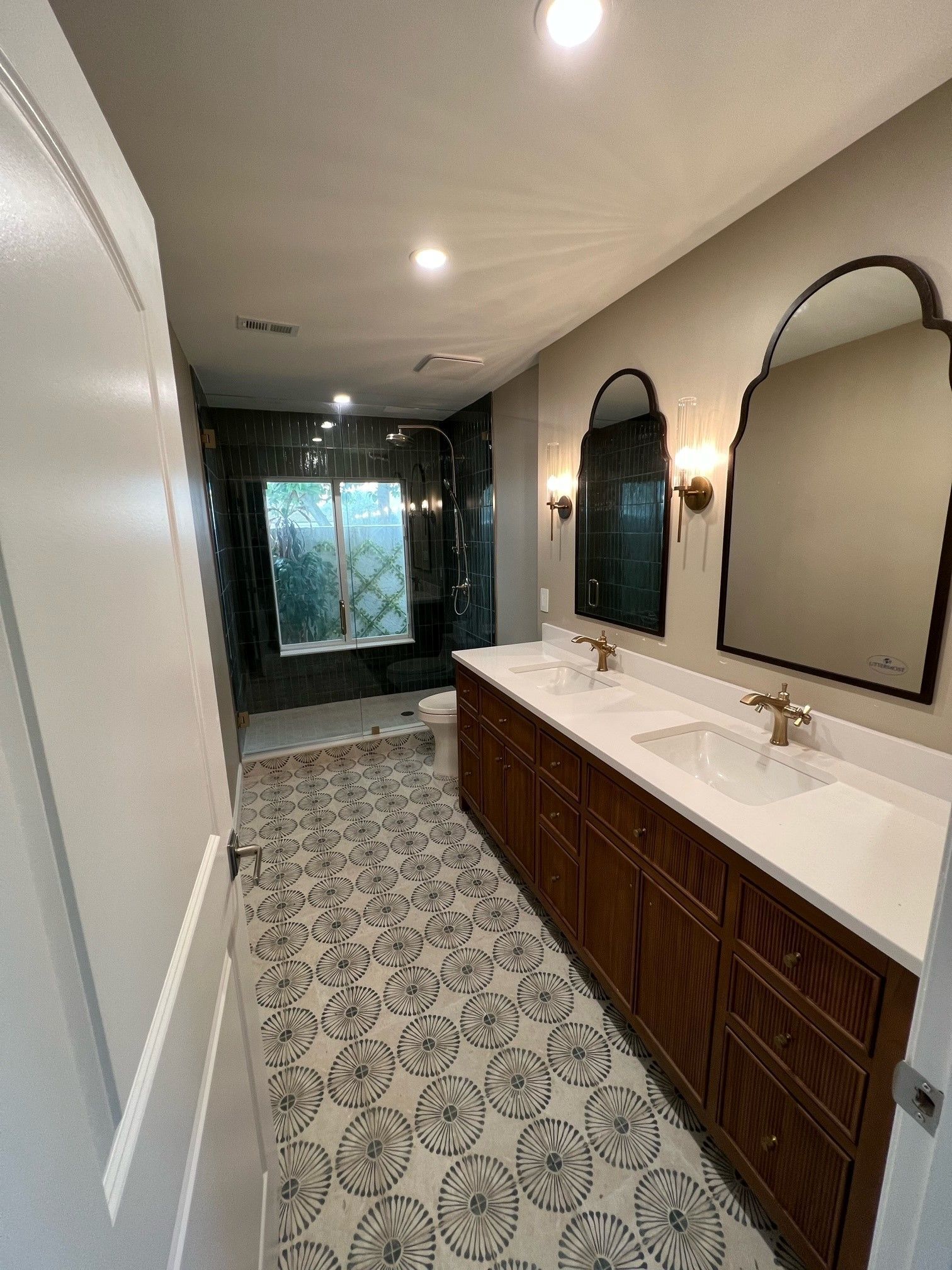 Bathroom with a long vanity, ornate mirrors, and patterned floor tile. Shower in the back.