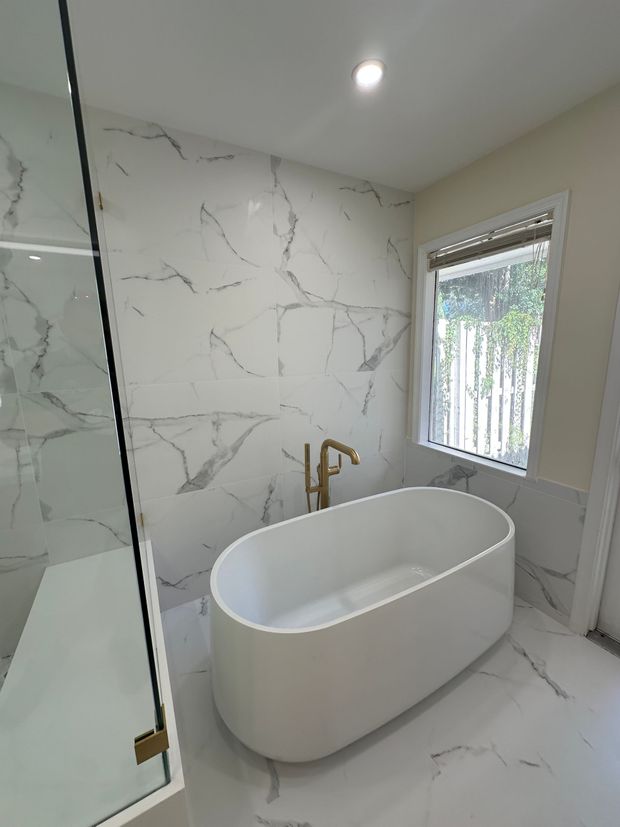 Bathroom with white marble tile, a modern tub, and gold faucet. Natural light streams in from a window.