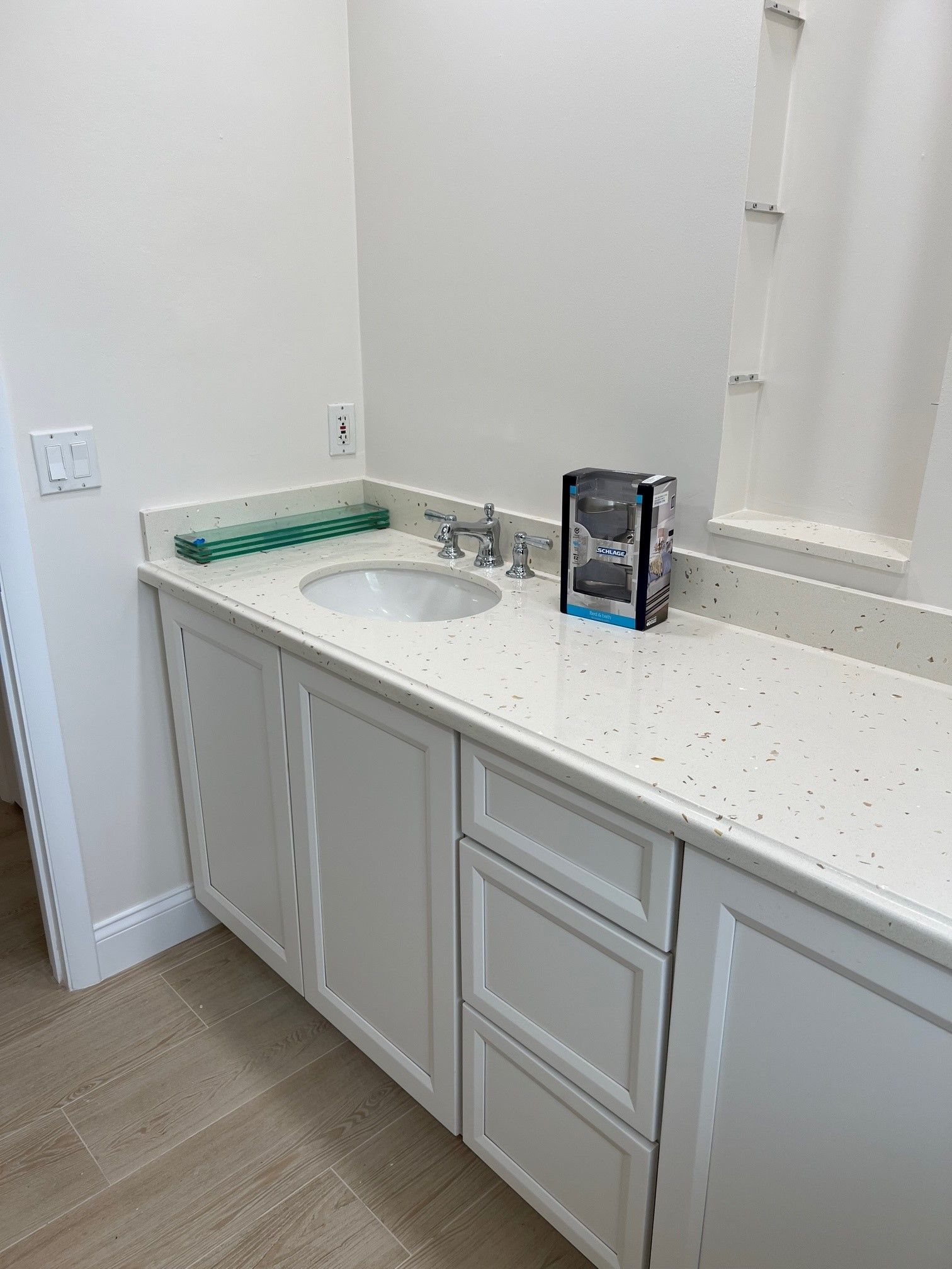 White bathroom vanity with sink, drawers, and speckled countertop; a green item and a black box sit on the counter.