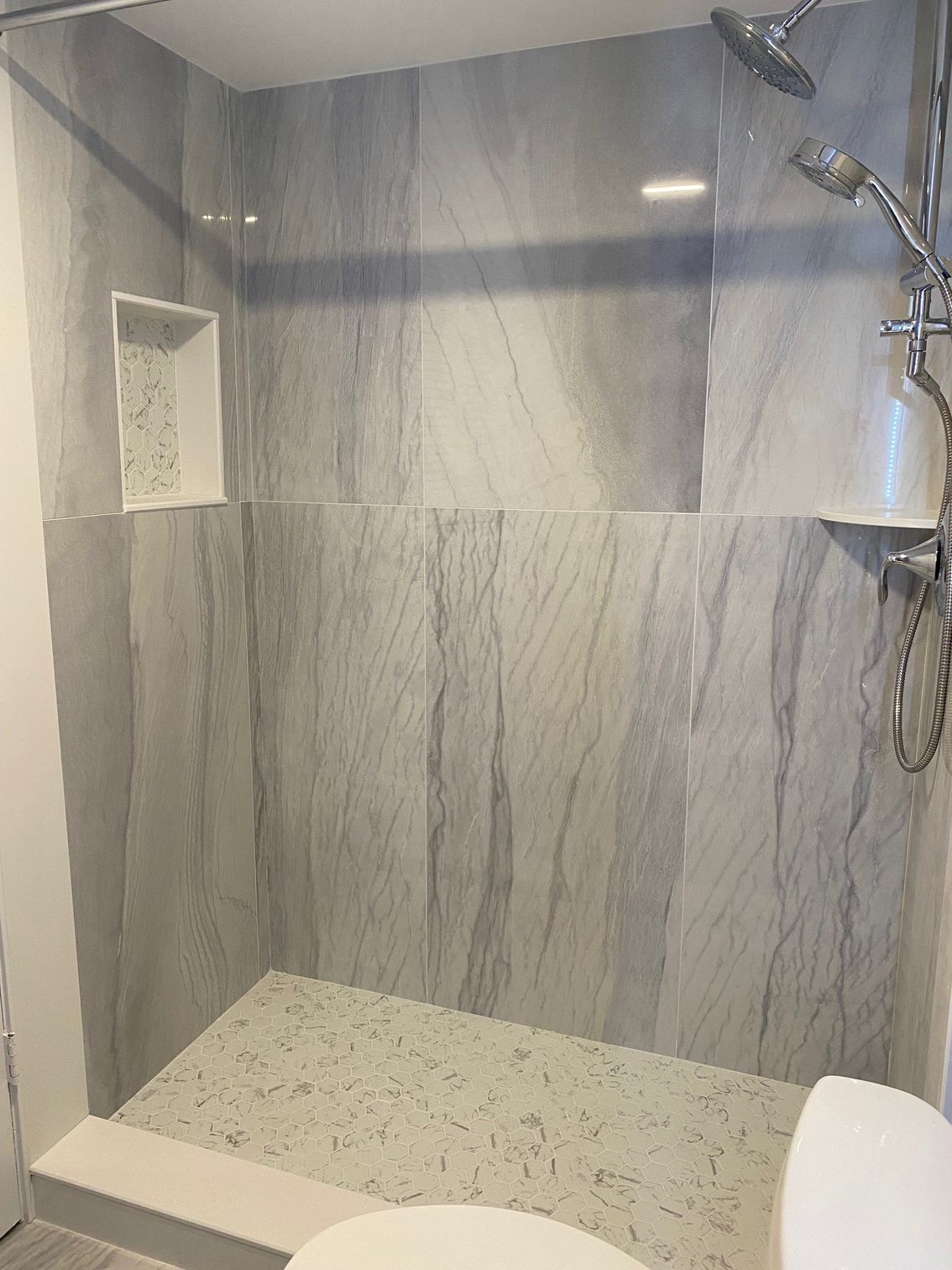 Shower stall with gray marbled tile walls and speckled floor. Showerhead and shelf visible.