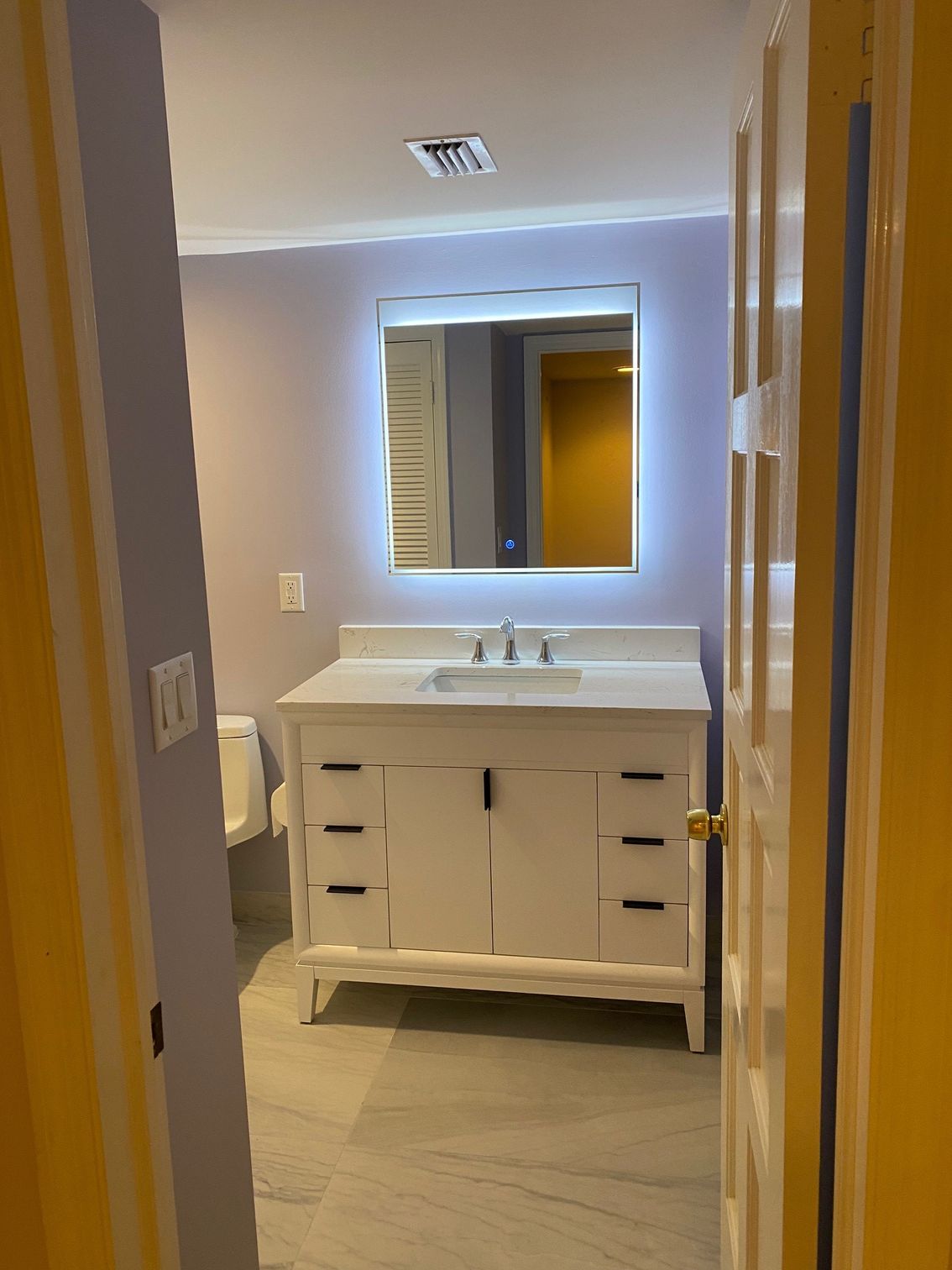 Bathroom with white vanity, lit mirror, light purple walls, and gray floor, viewed through an open door.