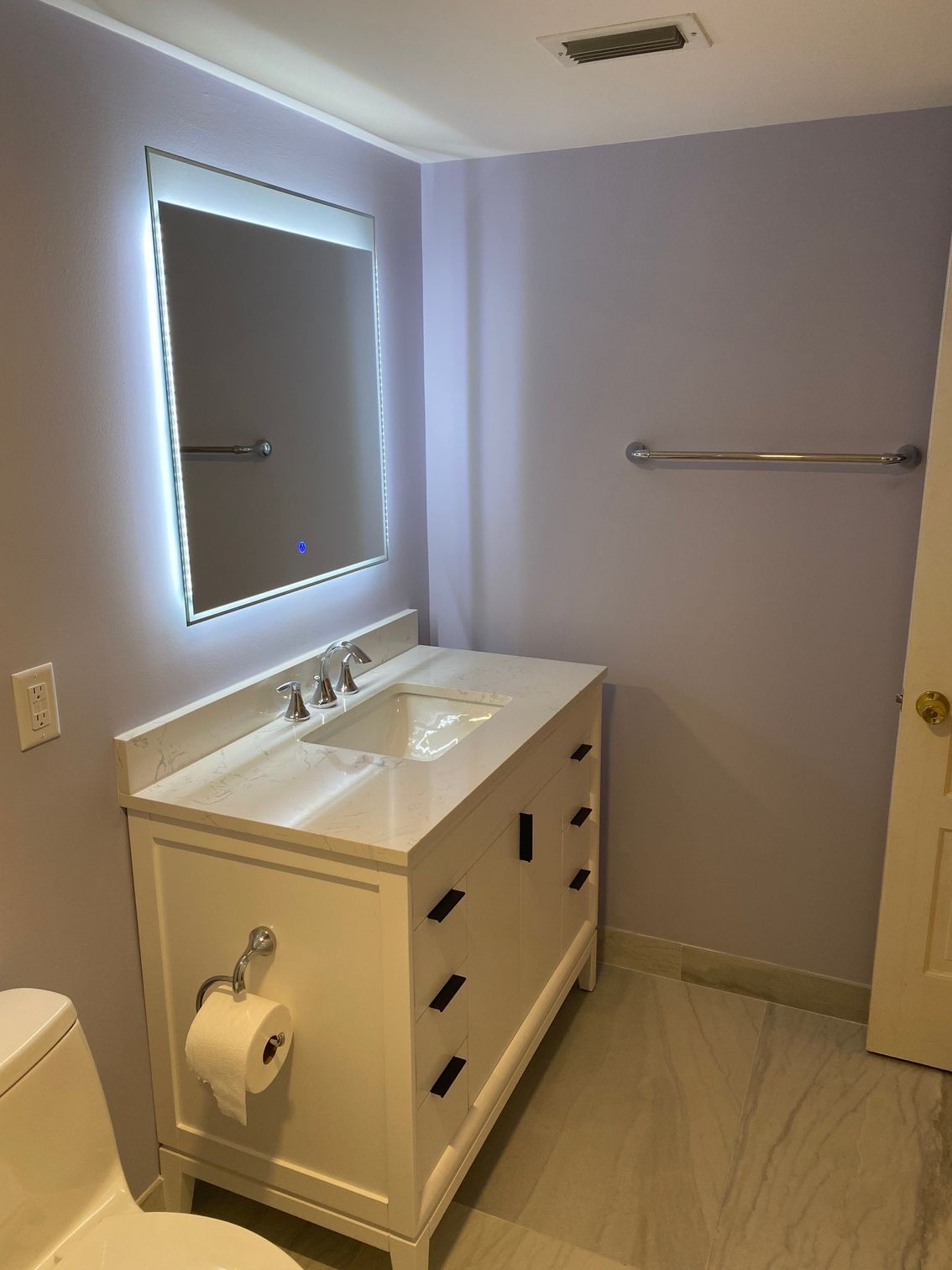White bathroom vanity with marble countertop, backlit mirror, and pale purple walls.