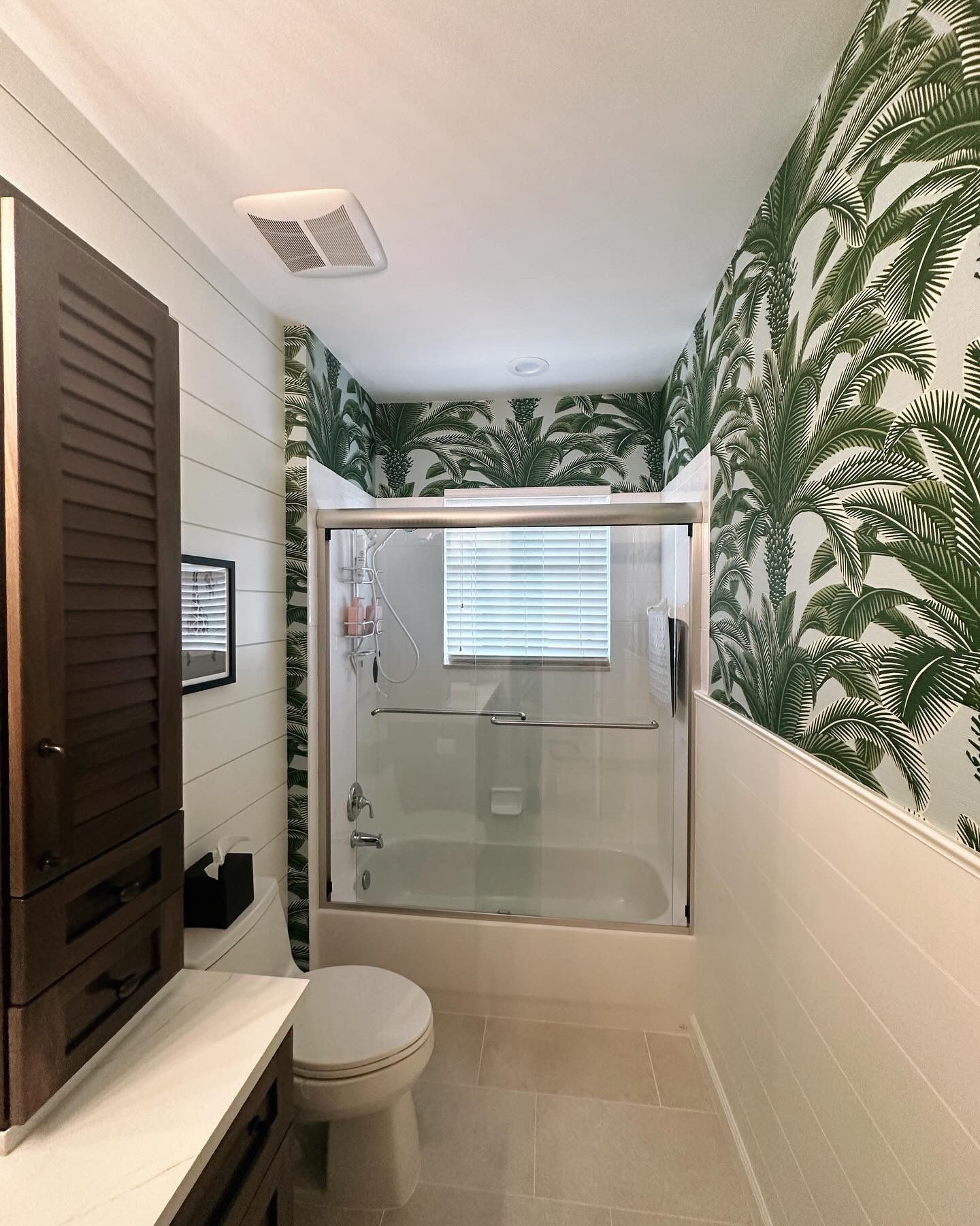 Bathroom with white shiplap wall, green palm wallpaper, glass shower, and wooden cabinet.