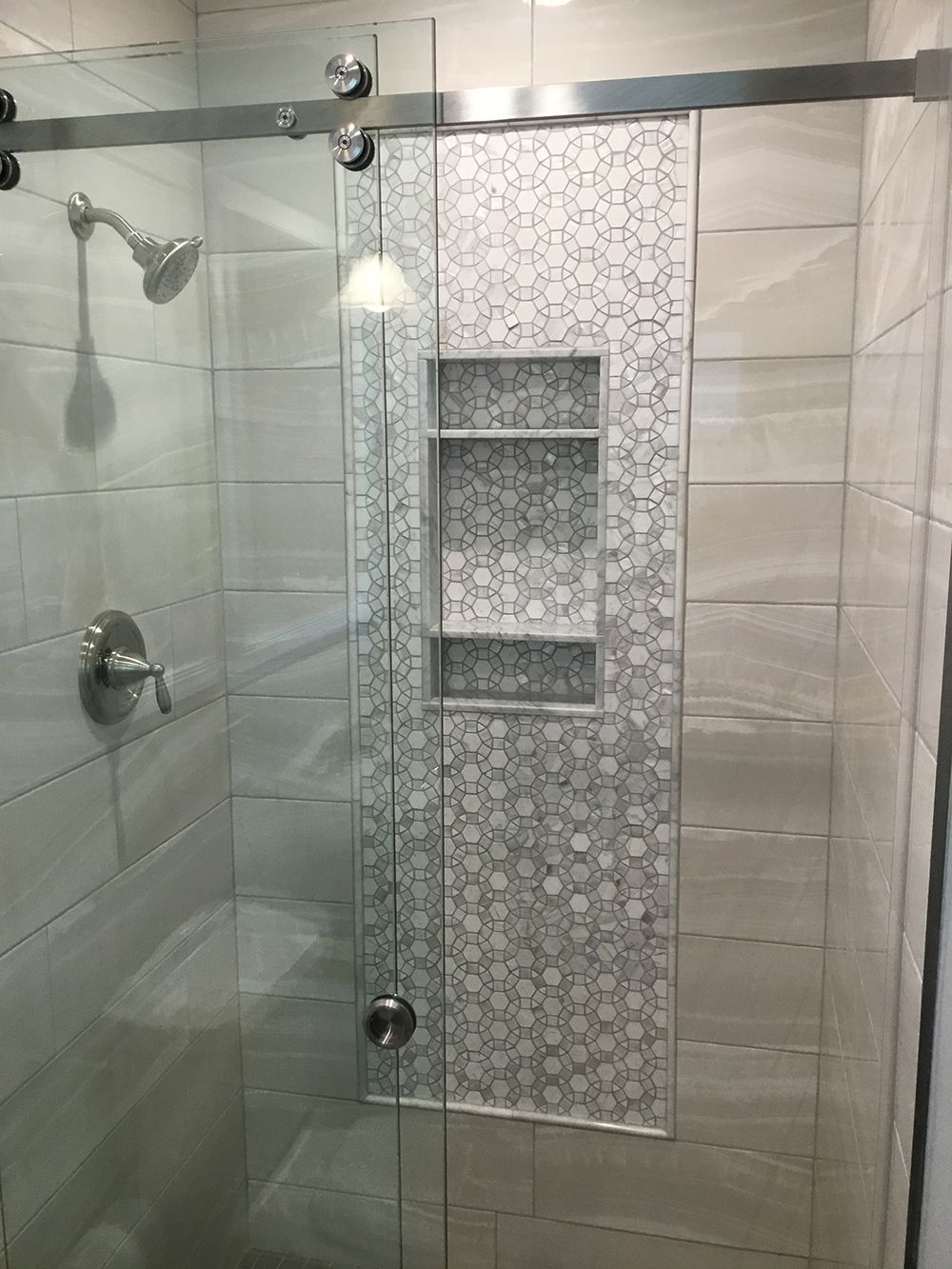 Shower with glass door, silver fixtures, built-in shelves with gray and white tile, light-colored walls.