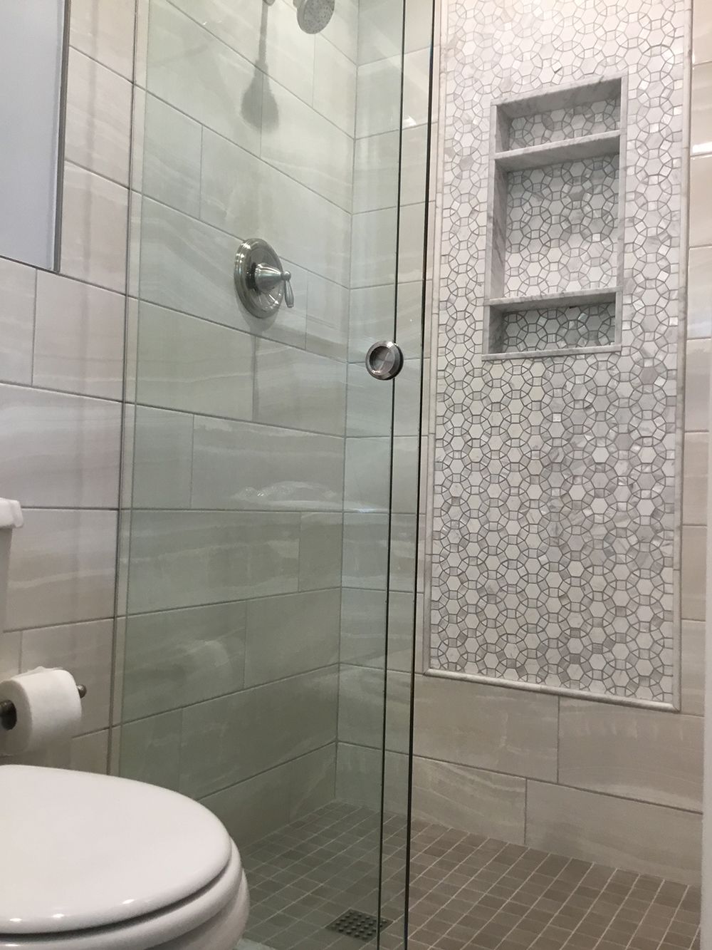 Bathroom with glass shower door, white tiled walls, toilet on the left, showerhead and faucet visible.