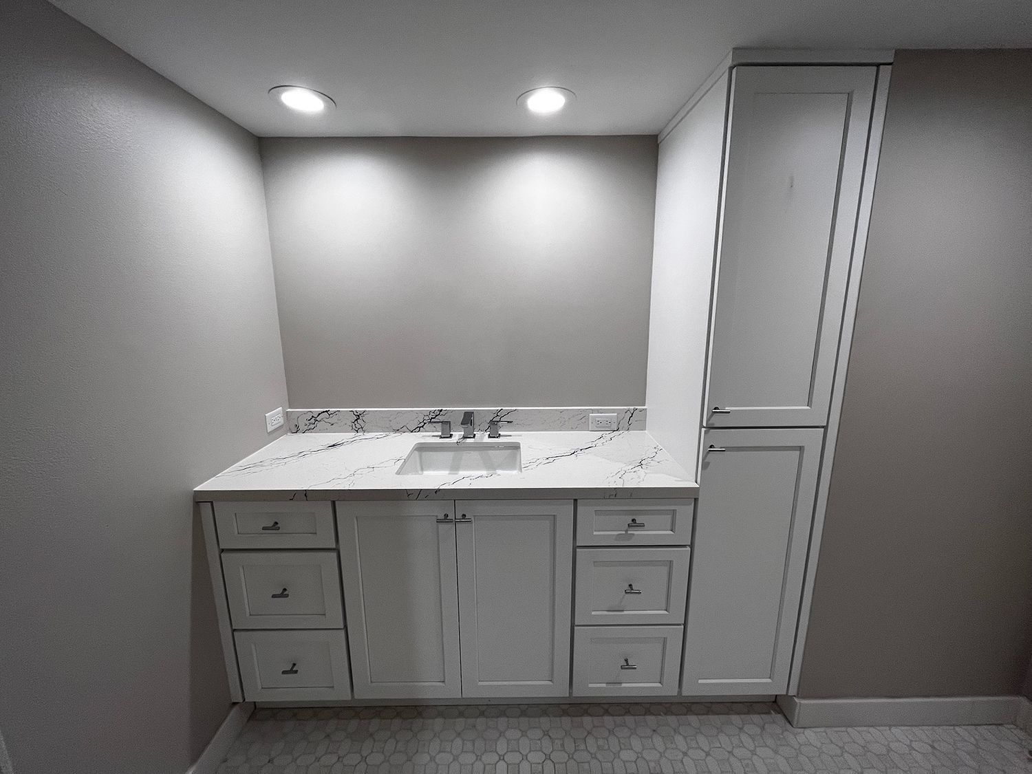 White bathroom vanity with countertop, sink, and cabinet. Gray walls, recessed lighting, and a tall cabinet to the right.