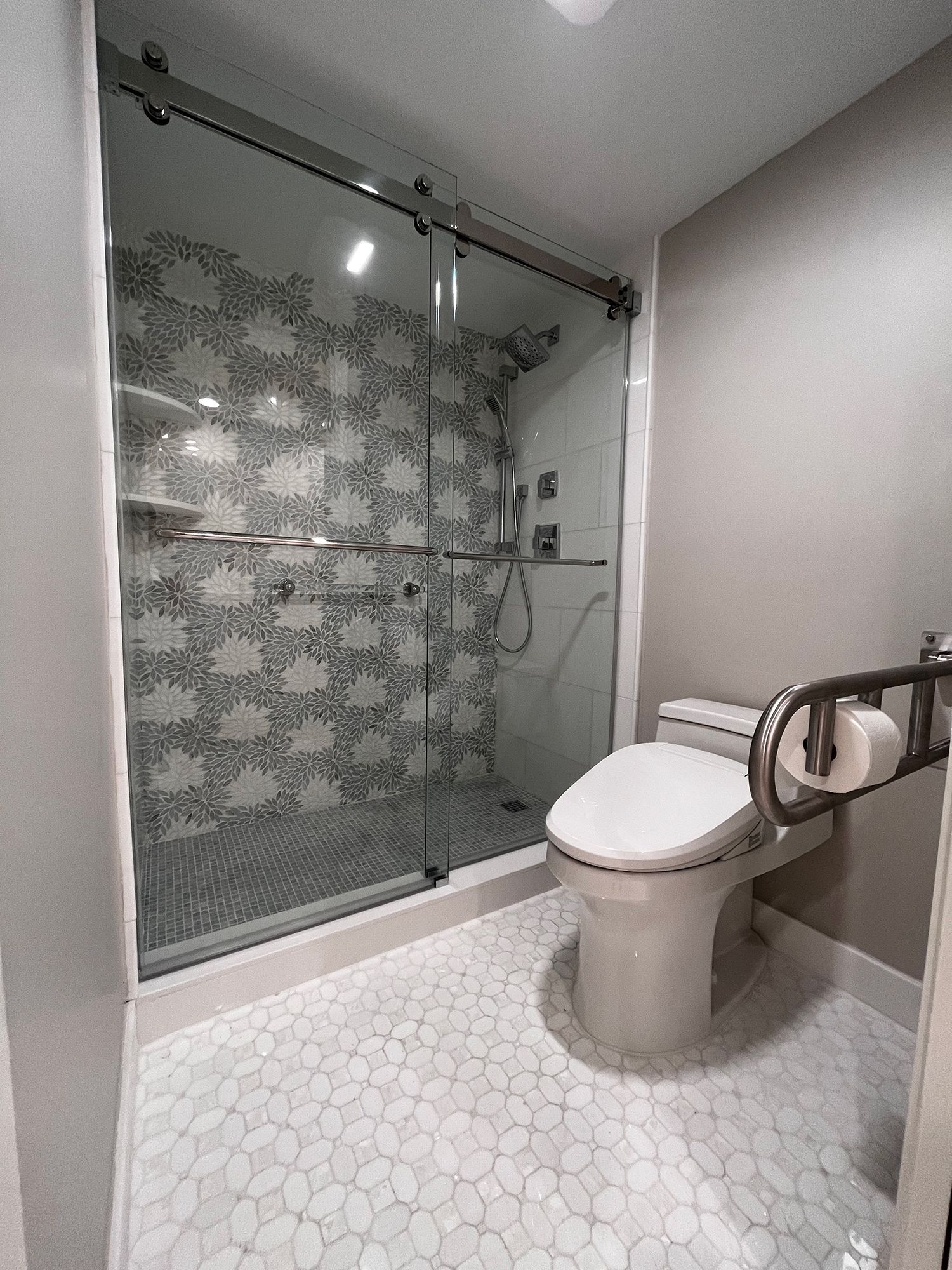 Bathroom with glass shower, patterned tile, pebble floor, and toilet with grab bar.