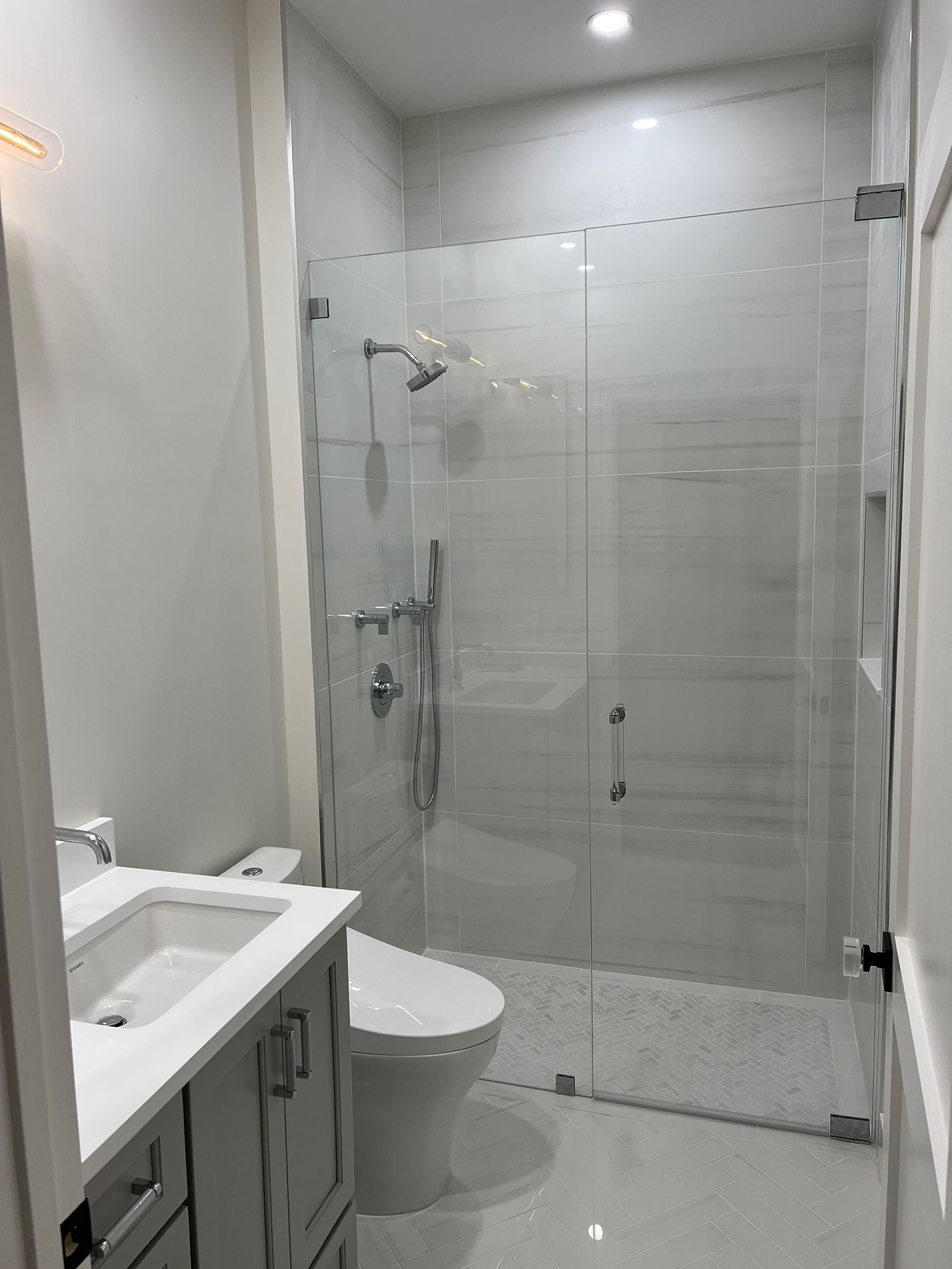 Modern bathroom with white tiled walls, glass shower, toilet, and vanity.
