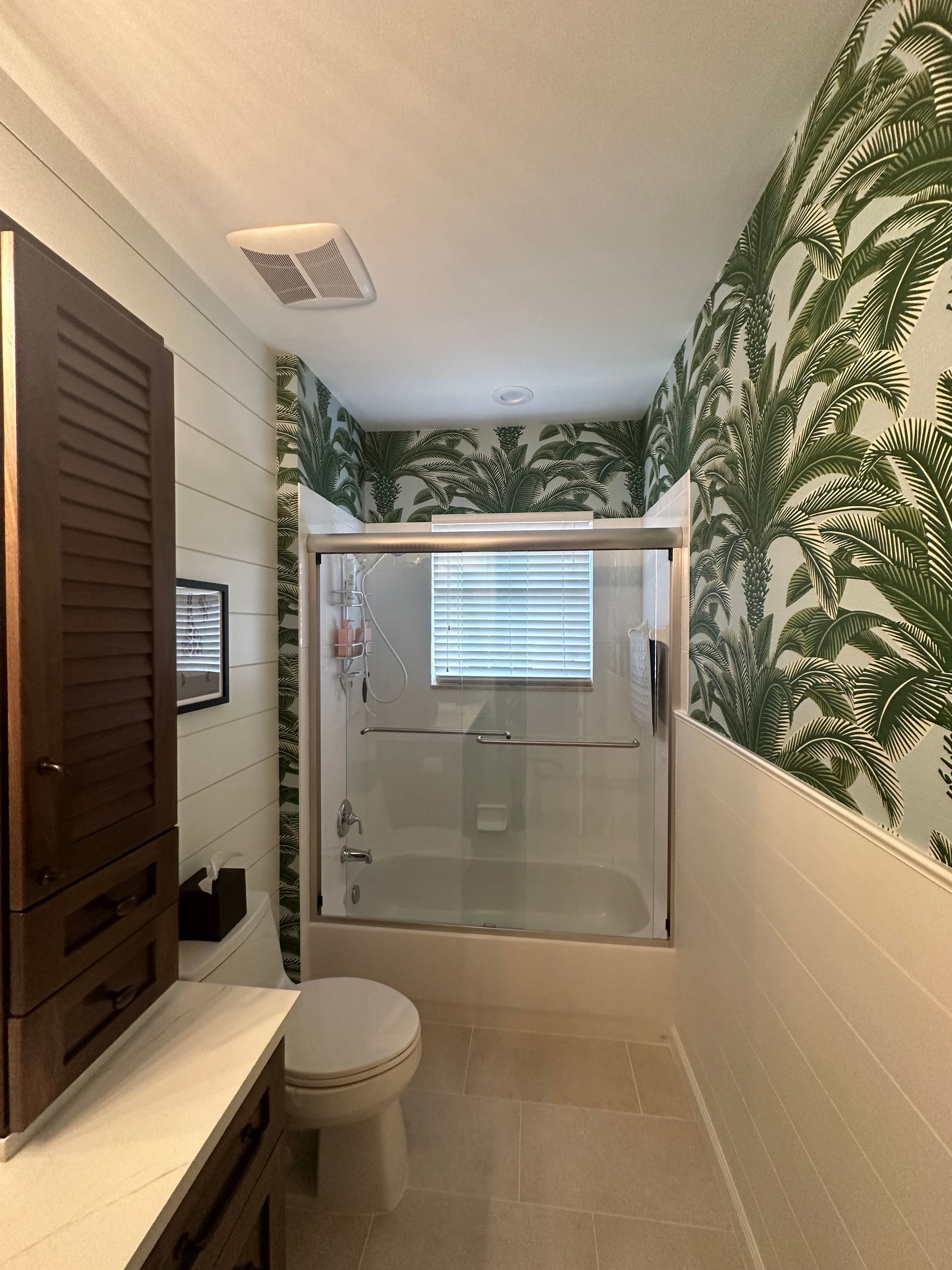 Bathroom with tropical palm tree wallpaper and white tile walls. Dark wood cabinet.