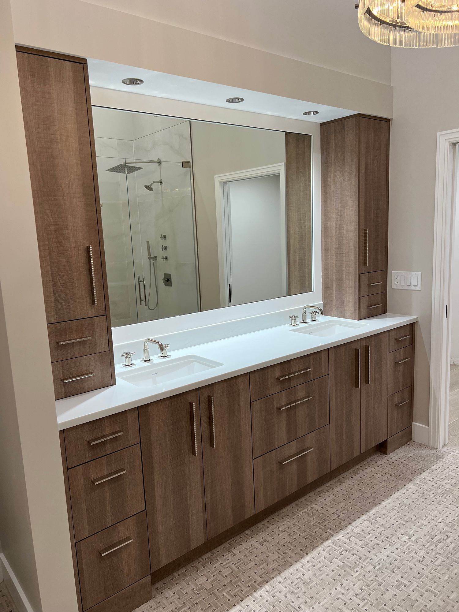 Bathroom with wood-grain cabinets, white countertops, large mirror, and a shower visible in the background.