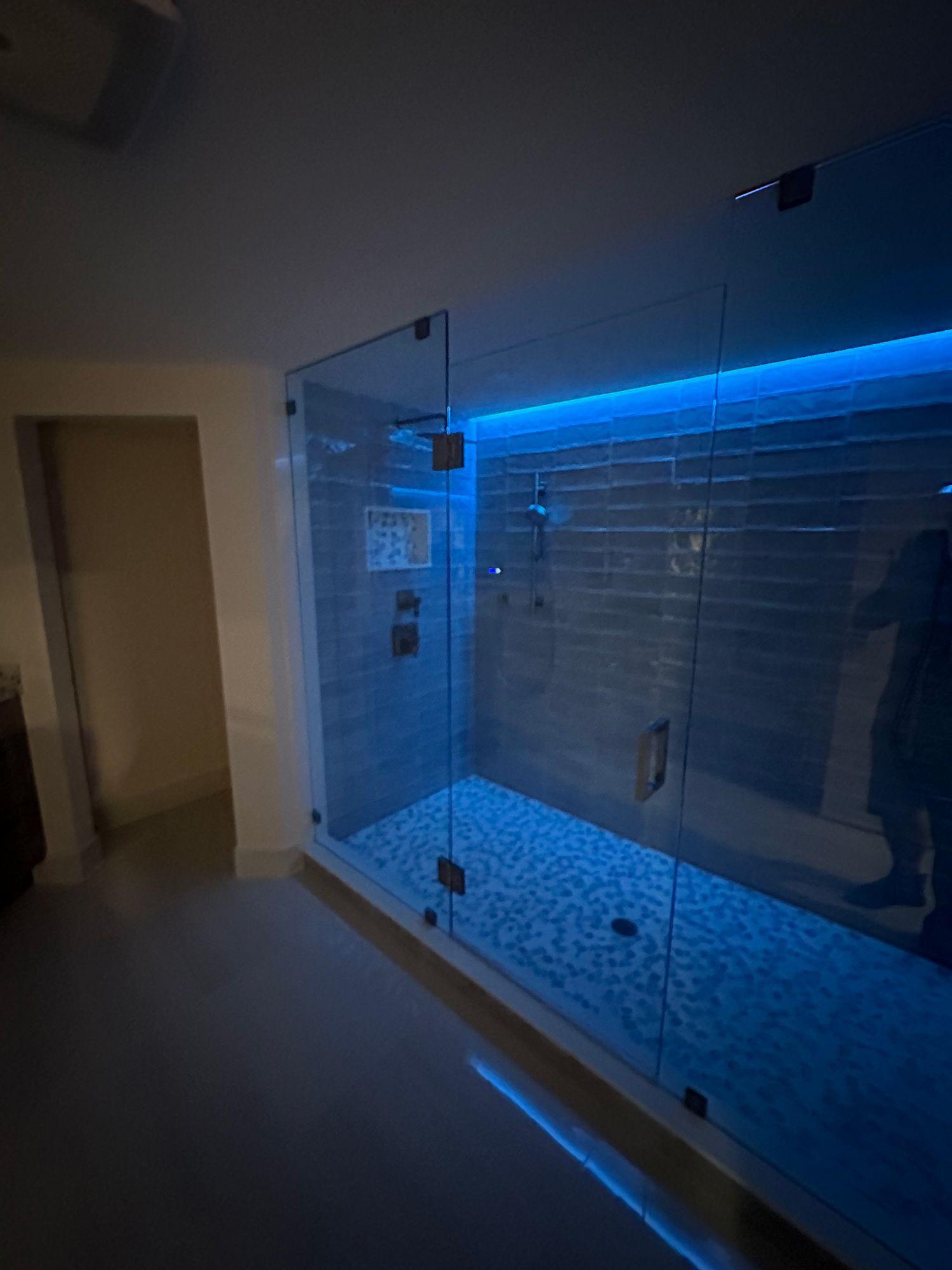 Glass-enclosed shower with blue LED lighting.  Mosaic tile floor, dark fixtures, and a glimpse of a doorway.
