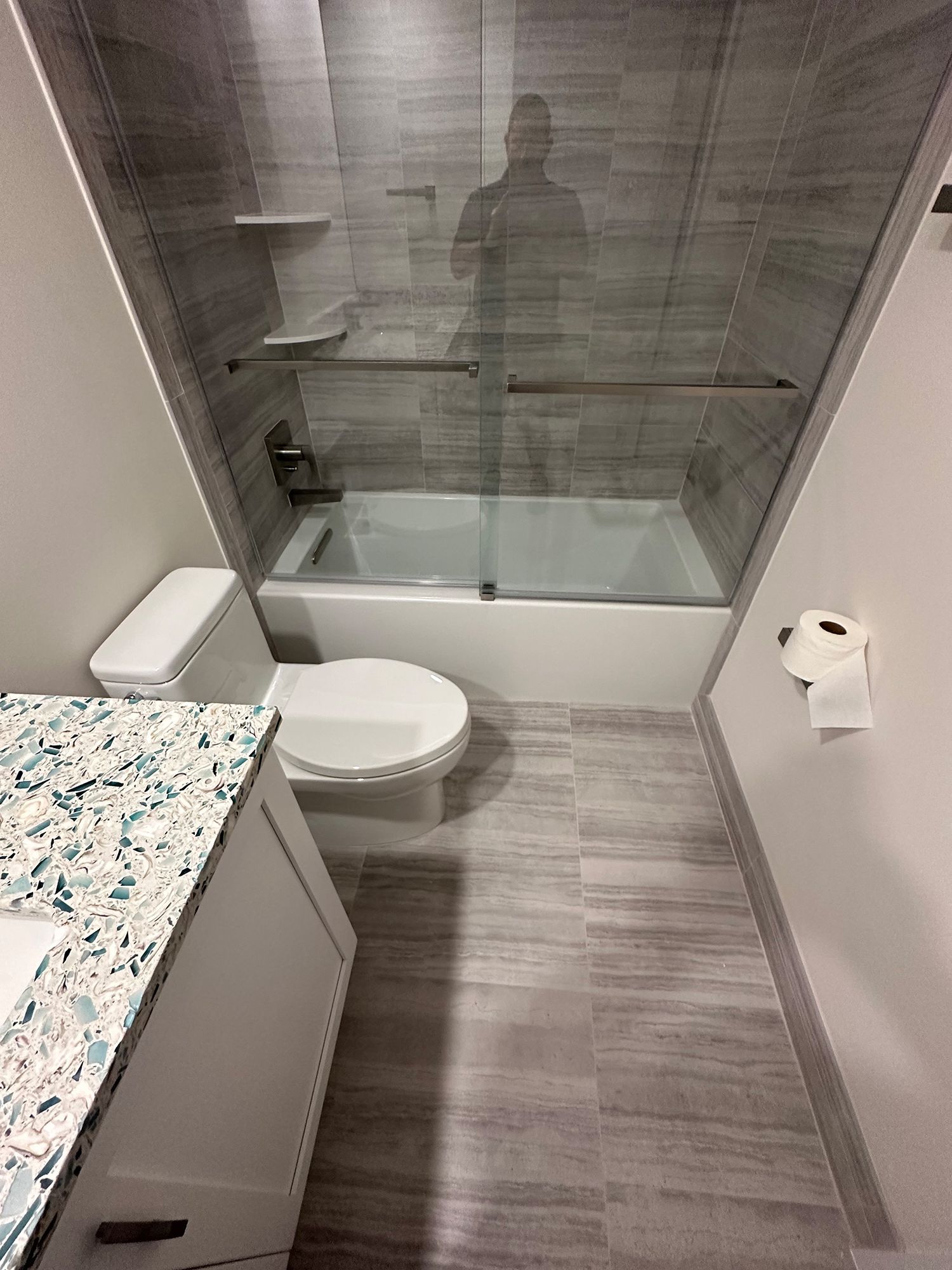 Bathroom with white toilet, tub, and vanity, gray tiled walls and floor. Person reflected in shower glass.
