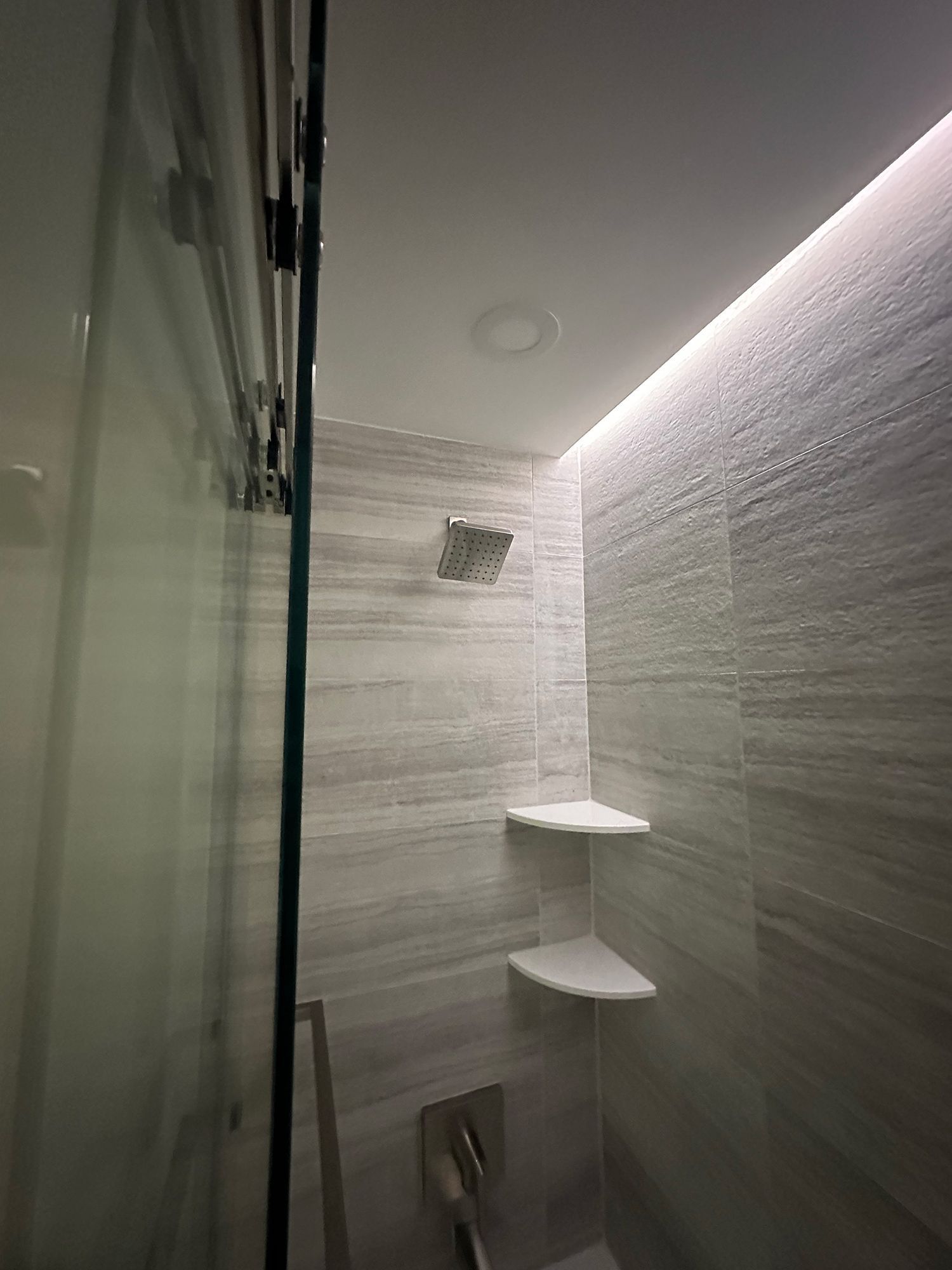 Shower with glass door, gray tile walls, corner shelves, and rain showerhead.