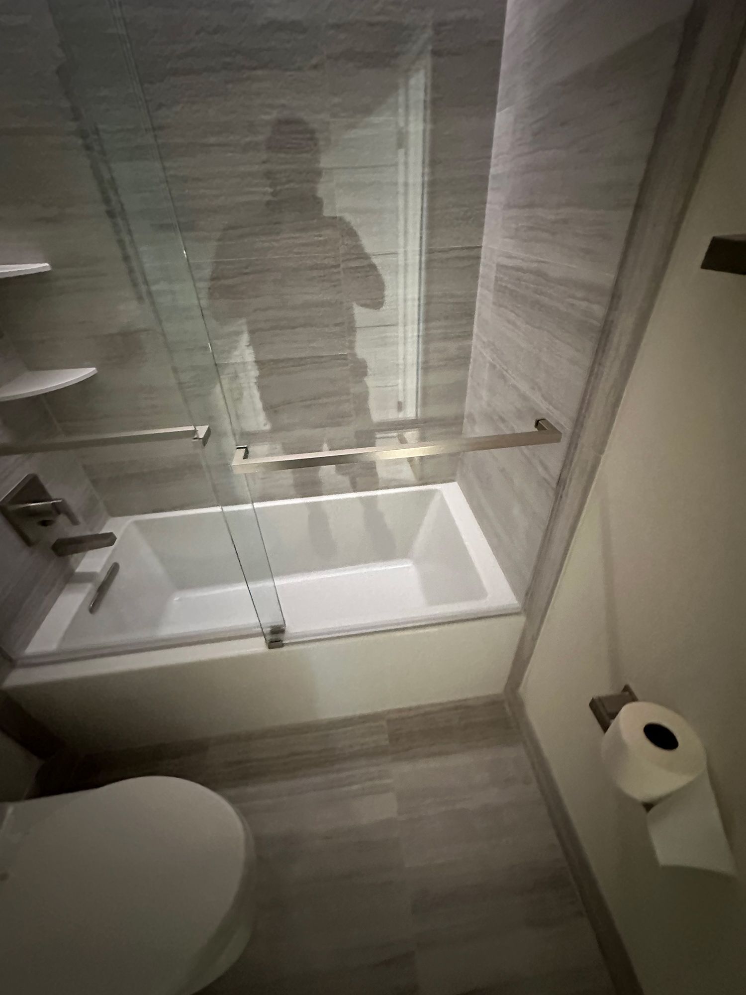 Bathroom with white tub, glass shower door, toilet, and gray tiled walls.