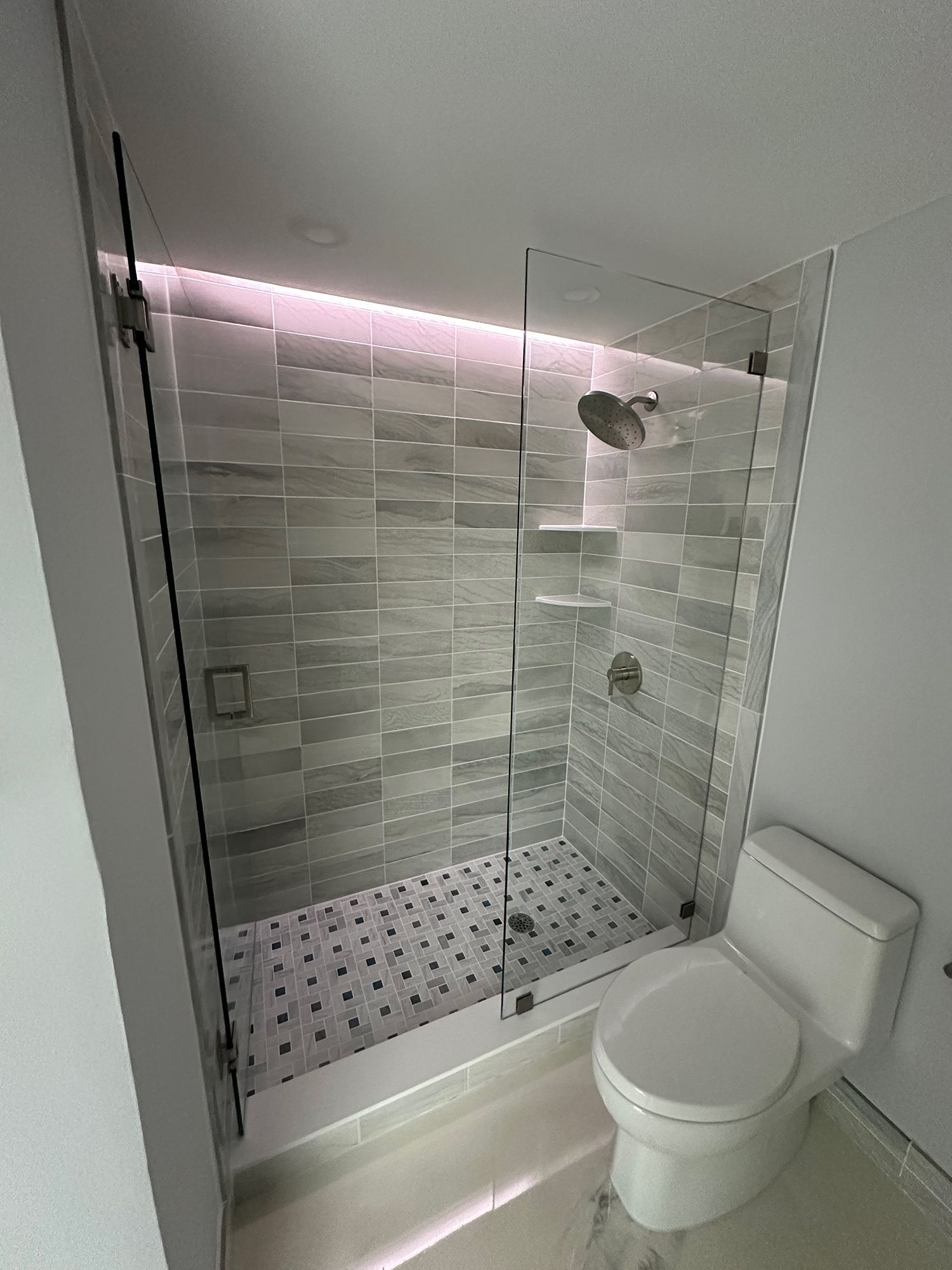 Modern shower with glass door and gray tiled walls, next to a white toilet.