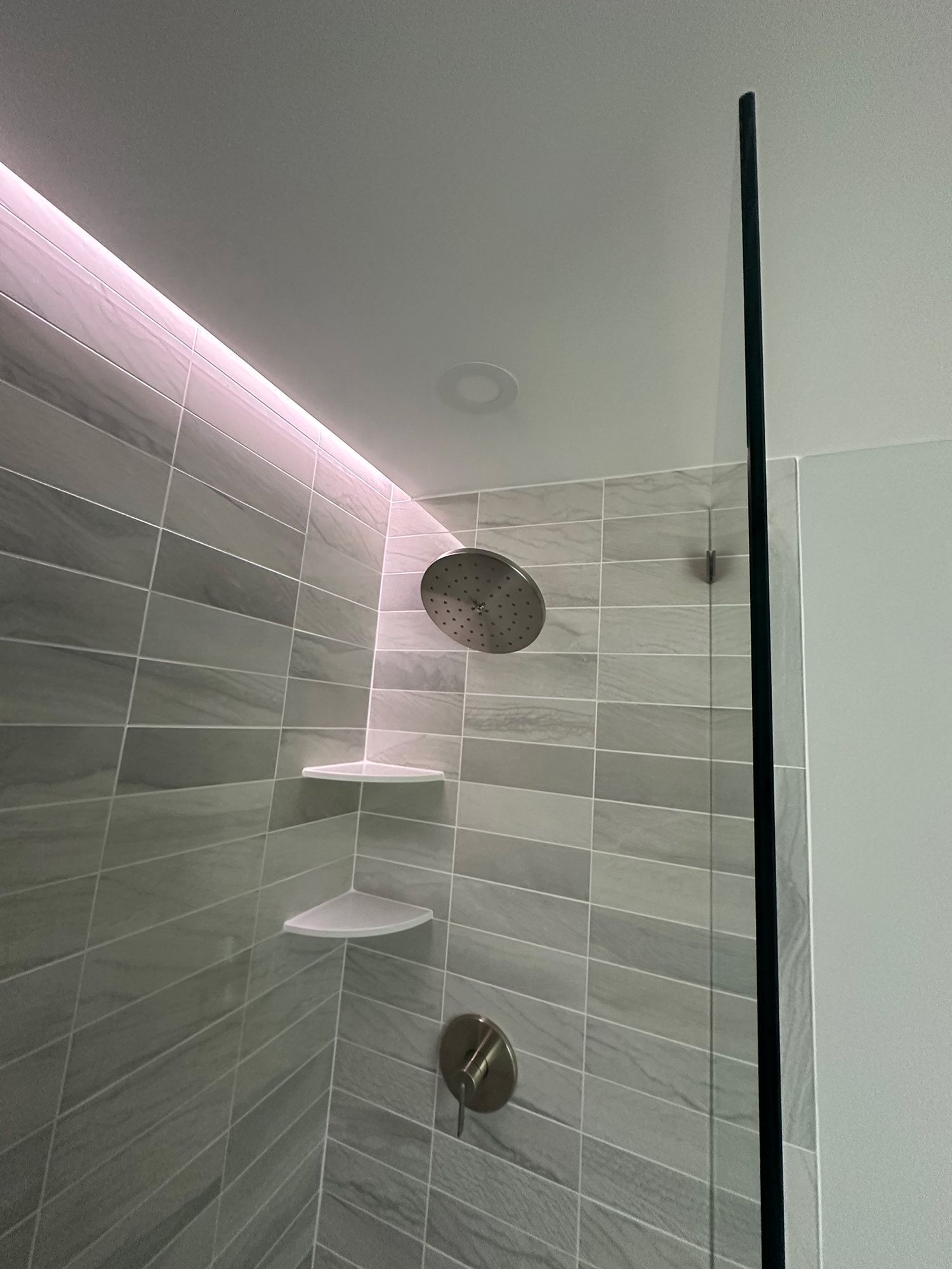 Shower with gray tiled walls, two circular shelves, and a glass door. A round showerhead and faucet are visible.