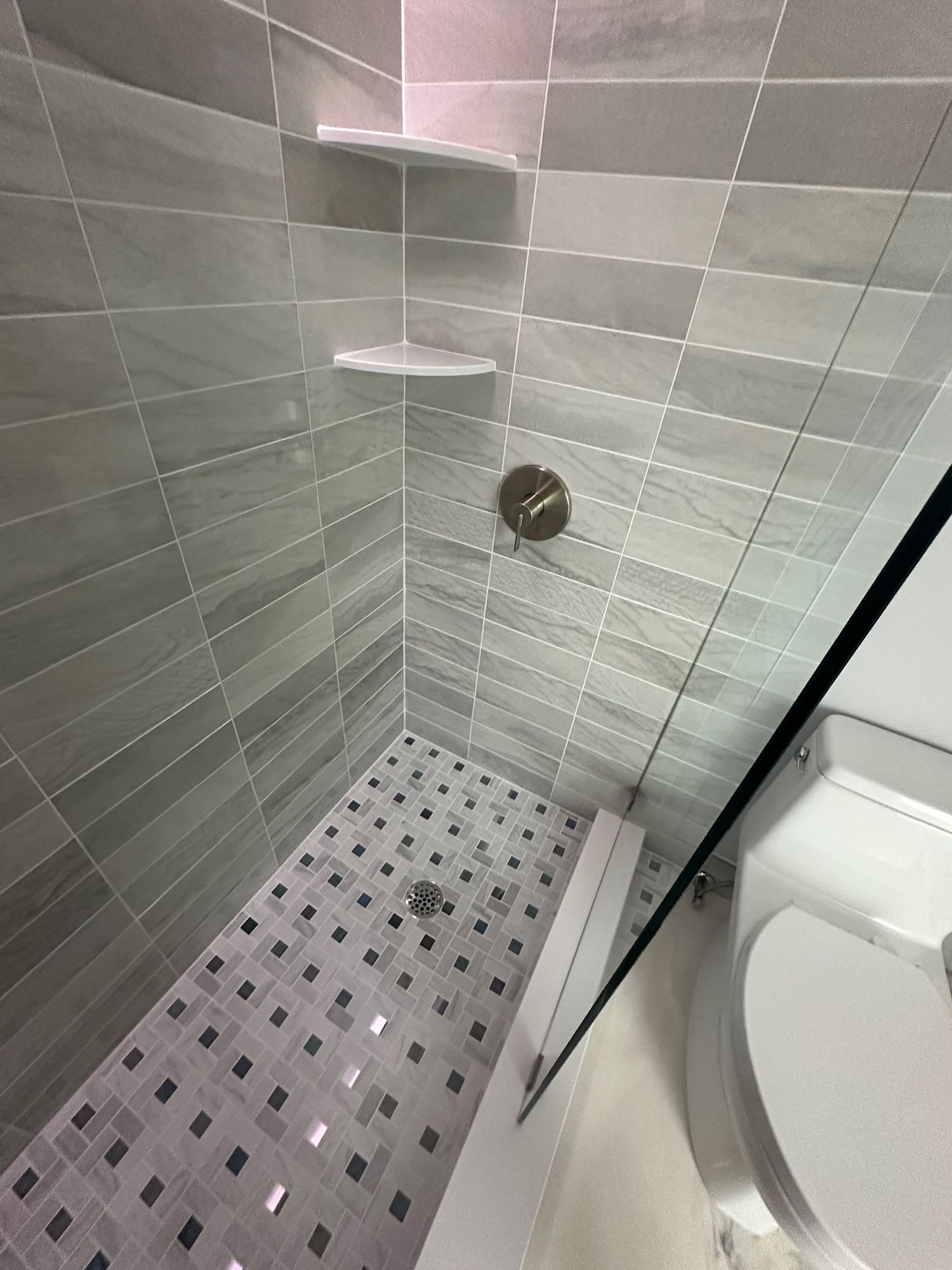 Shower with gray tile walls, corner shelves, and mosaic floor. Toilet visible on the right.