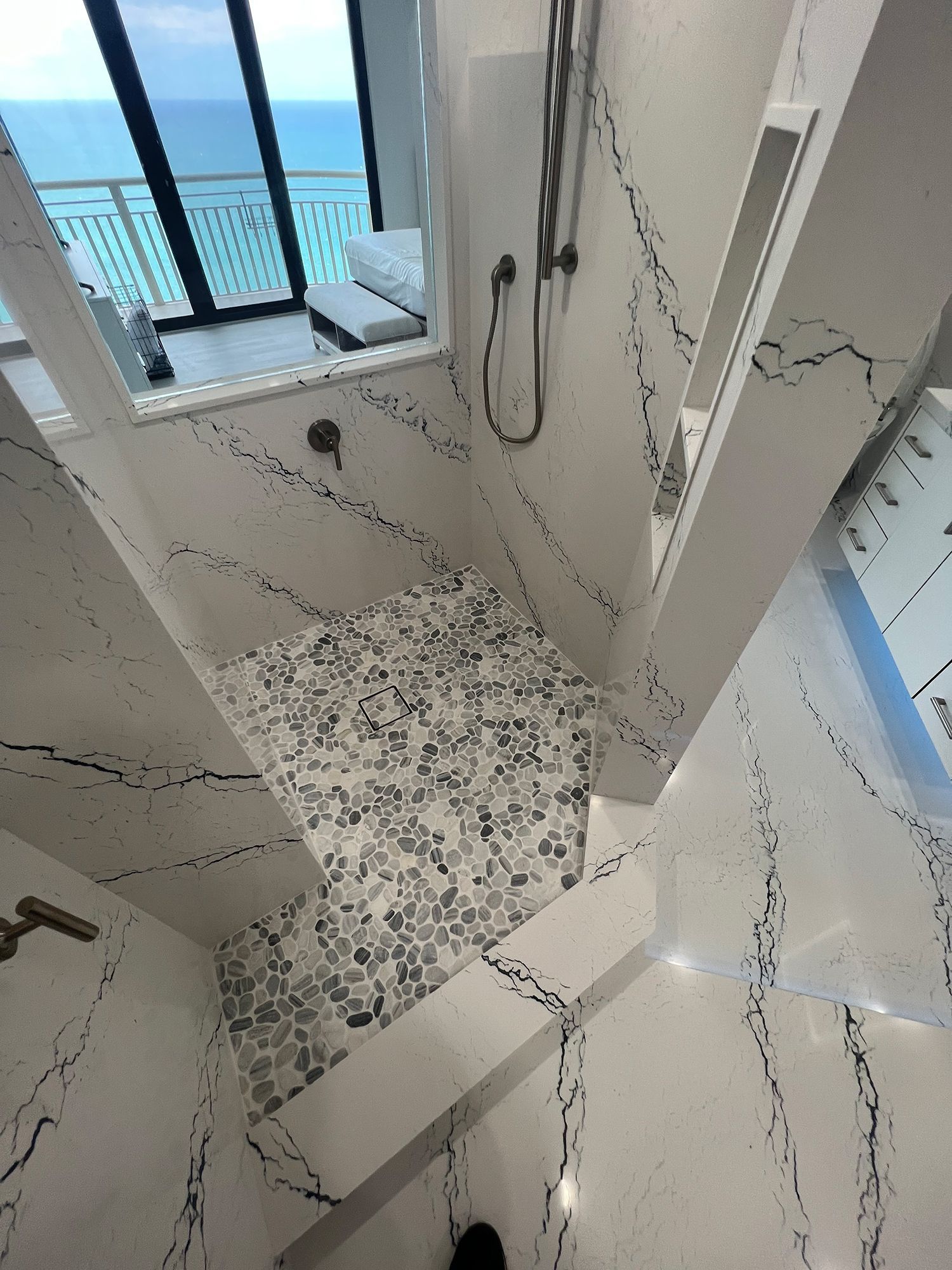 Luxurious shower with ocean view: White marble, pebble floor, glass enclosure, and modern fixtures.