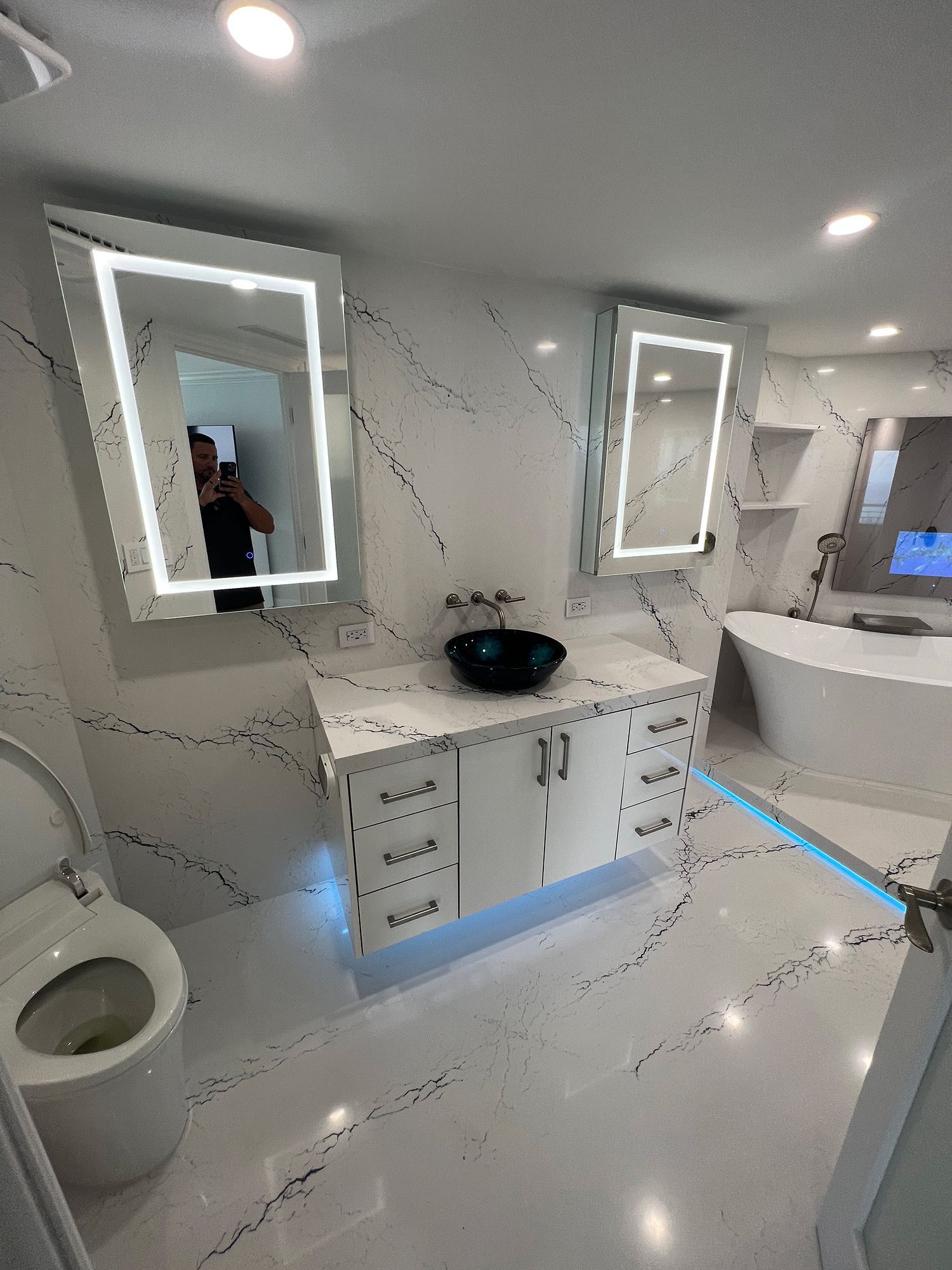 Modern bathroom with white marble-look surfaces, two backlit mirrors, black sink, and a white soaking tub.