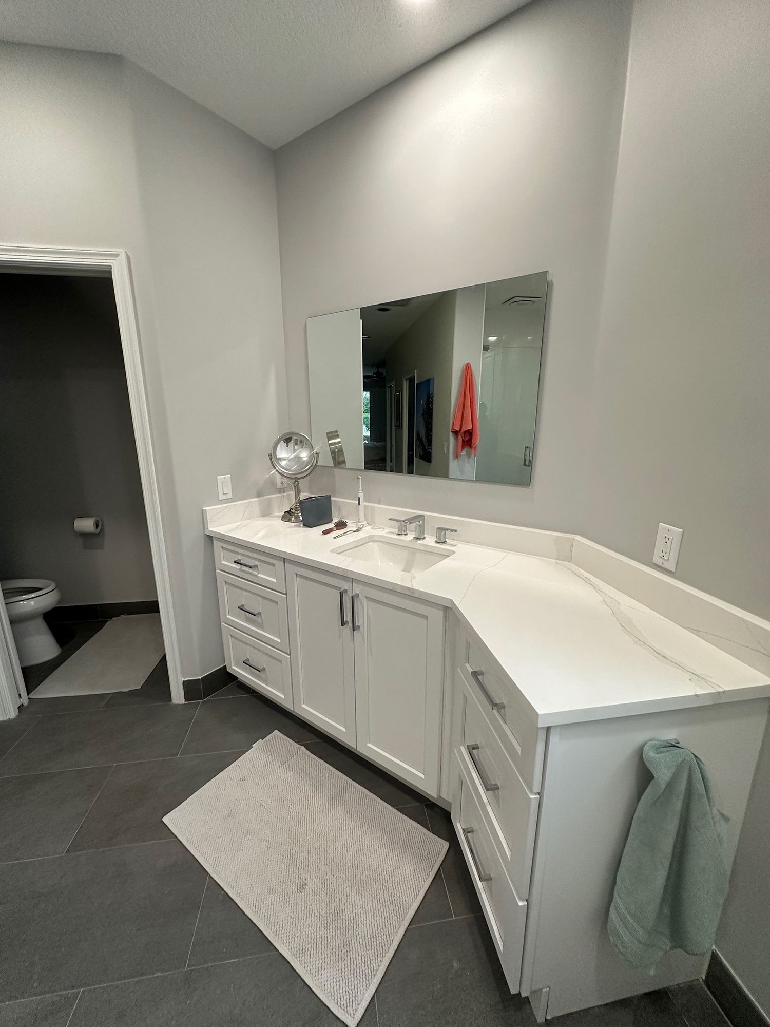 Bathroom with white vanity, large mirror, and gray walls. Towel on side, toilet visible in doorway.