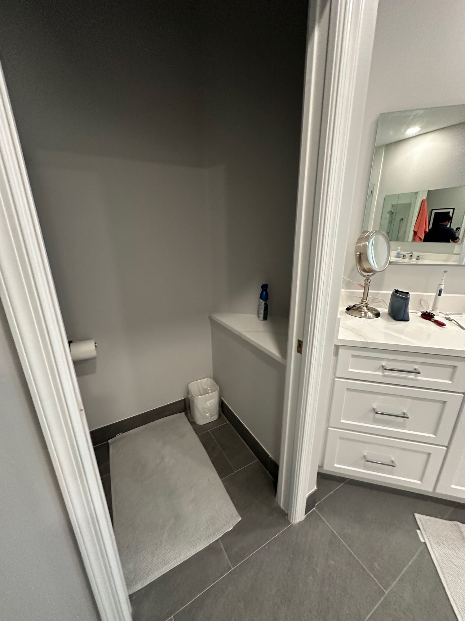 Bathroom with a toilet in a small alcove. A white cabinet and mirror are visible on the right. Gray tile floor.