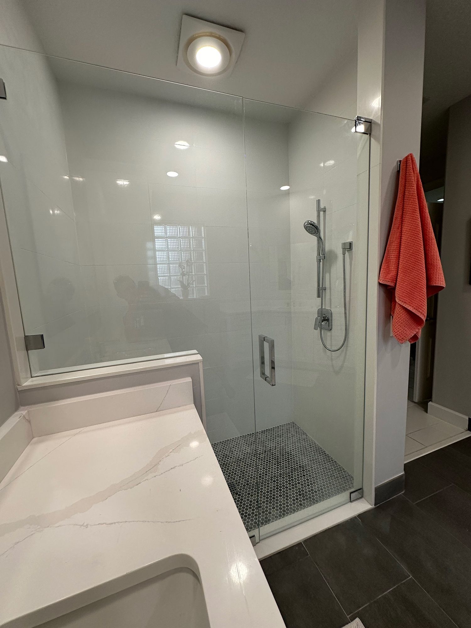 Bathroom with white countertop, glass shower, gray floor, and orange towel.