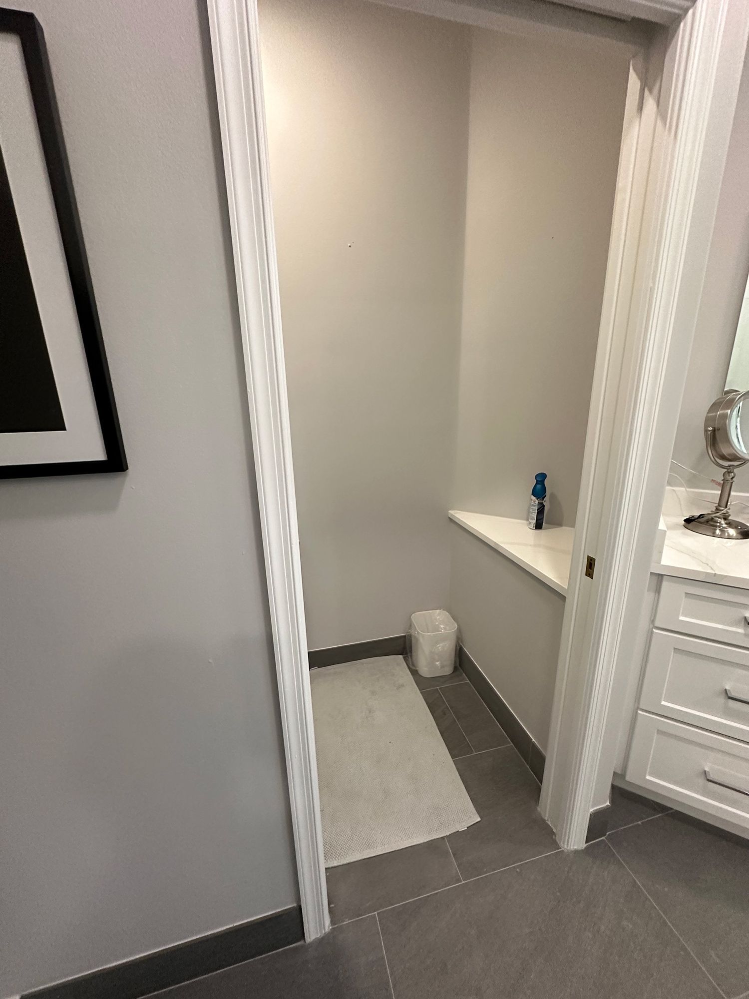 Bathroom with a gray floor, white walls, and a small built-in shelf. A white rug and trash can are in the space.