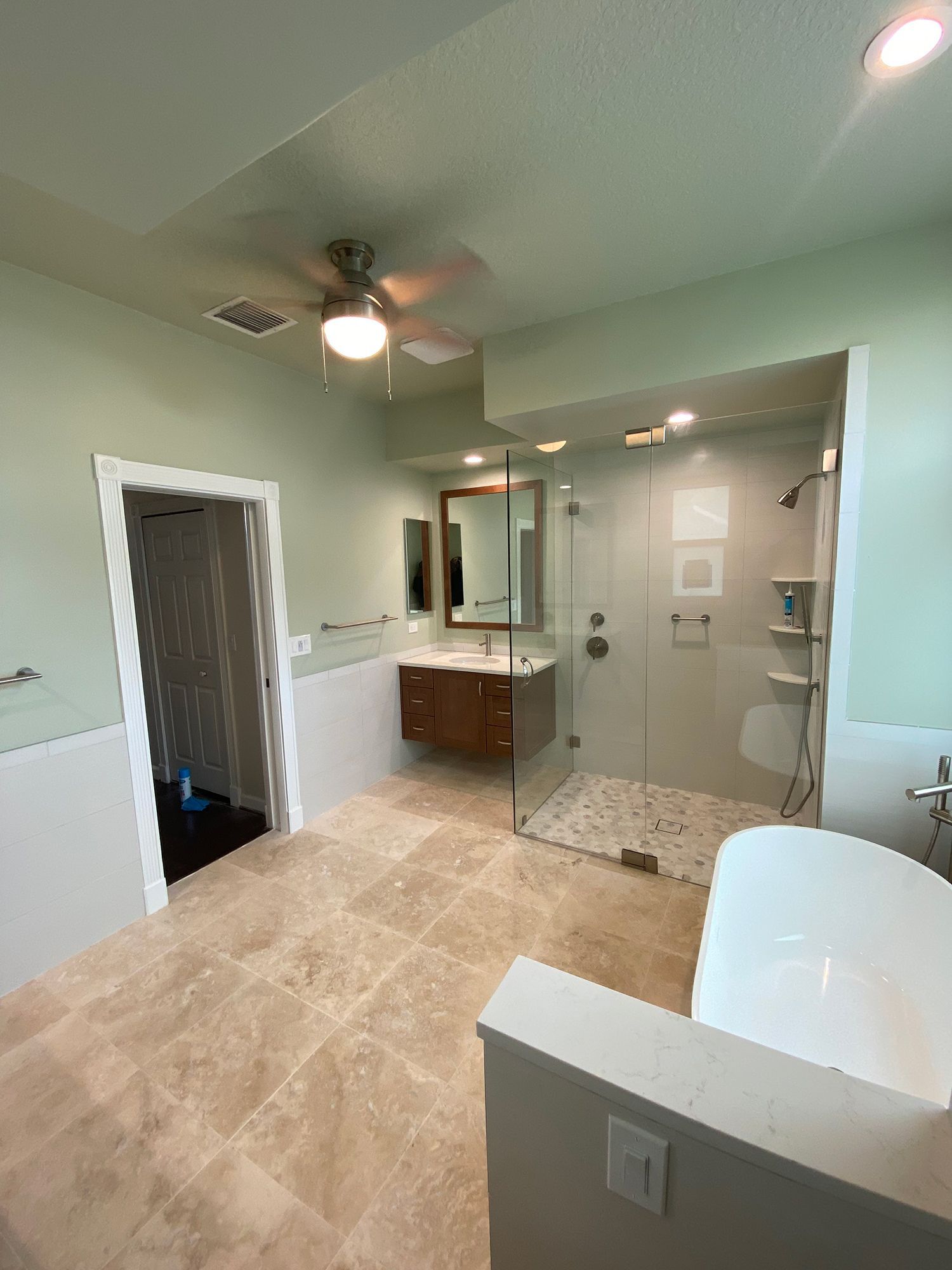 Spacious bathroom with glass shower, freestanding tub, and a vanity. Light green walls, stone tile floor.