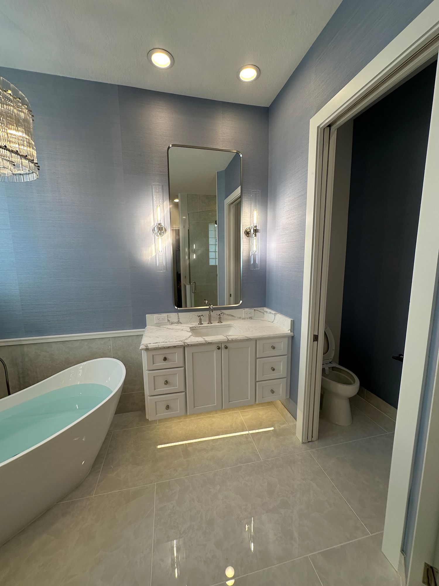 Bathroom with blue walls, white vanity, large mirror, soaking tub, and open doorway to a toilet area.
