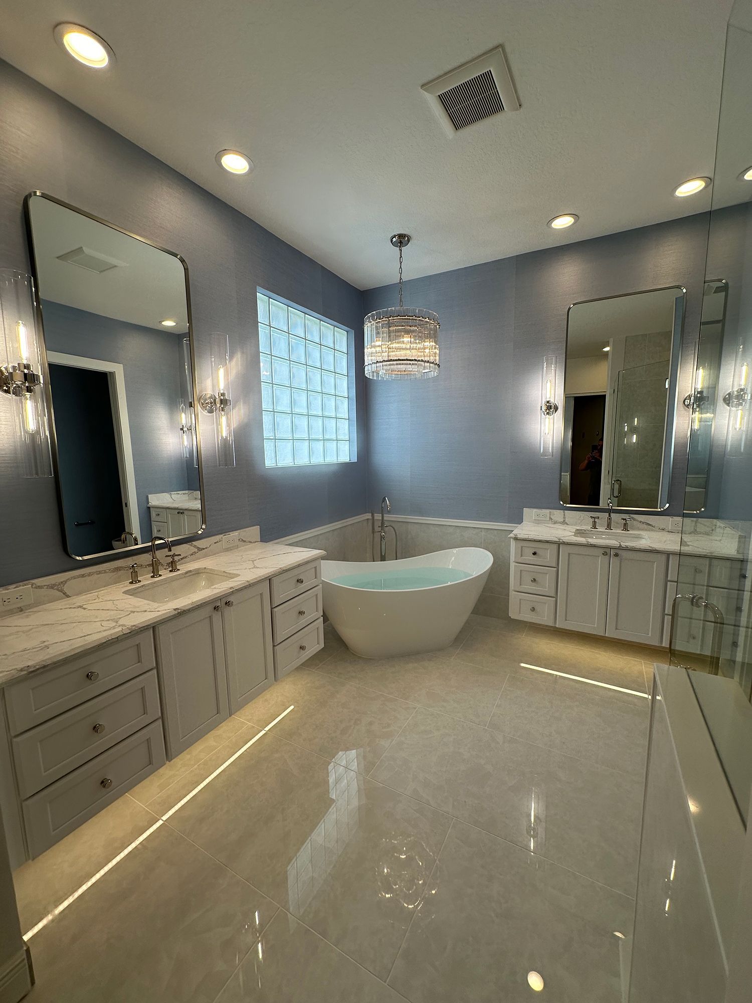 Modern bathroom with blue walls, white cabinets, freestanding tub, and large mirrors.