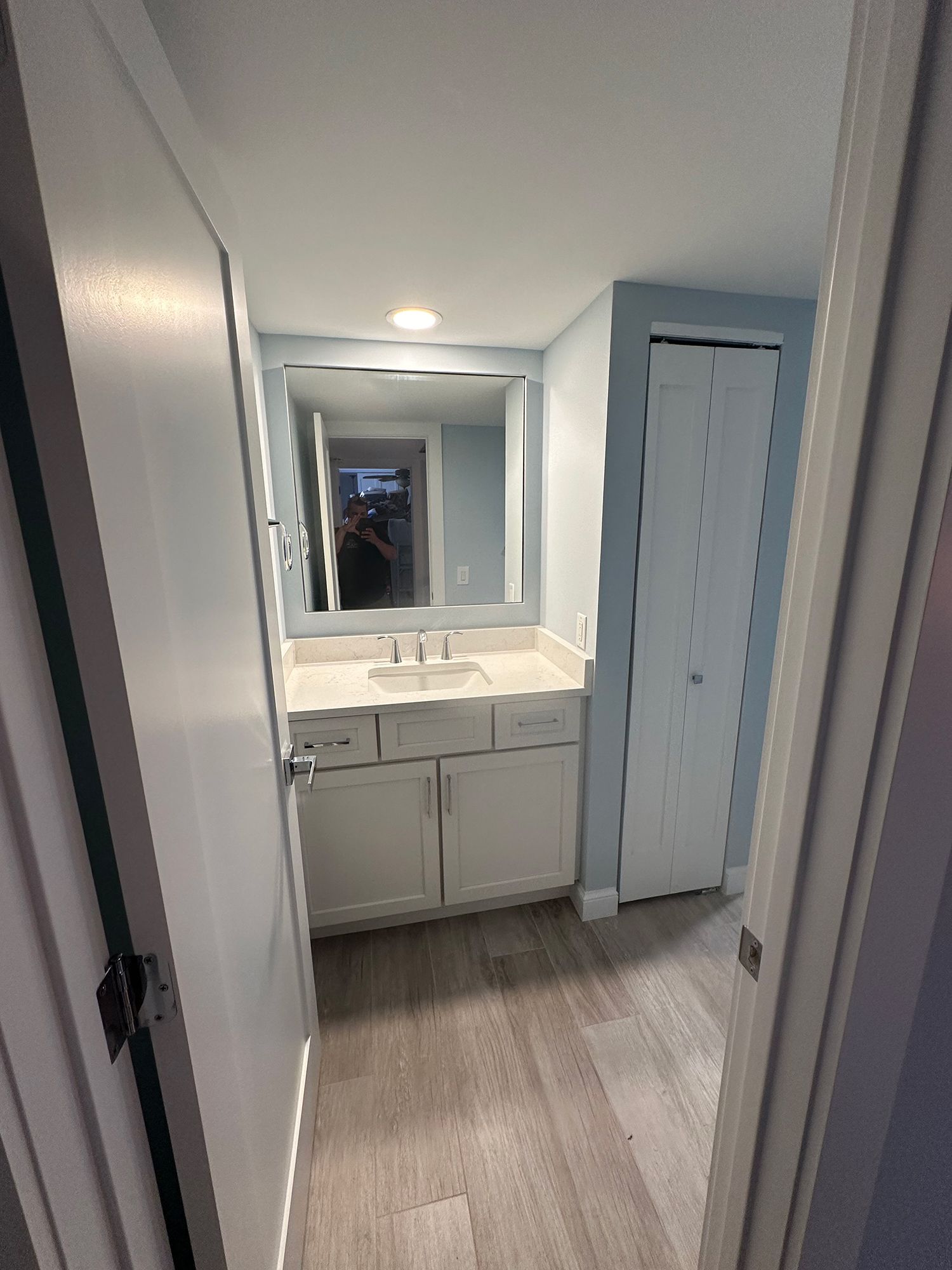 Bathroom with white vanity, large mirror, and closed closet door; light blue walls, gray flooring.