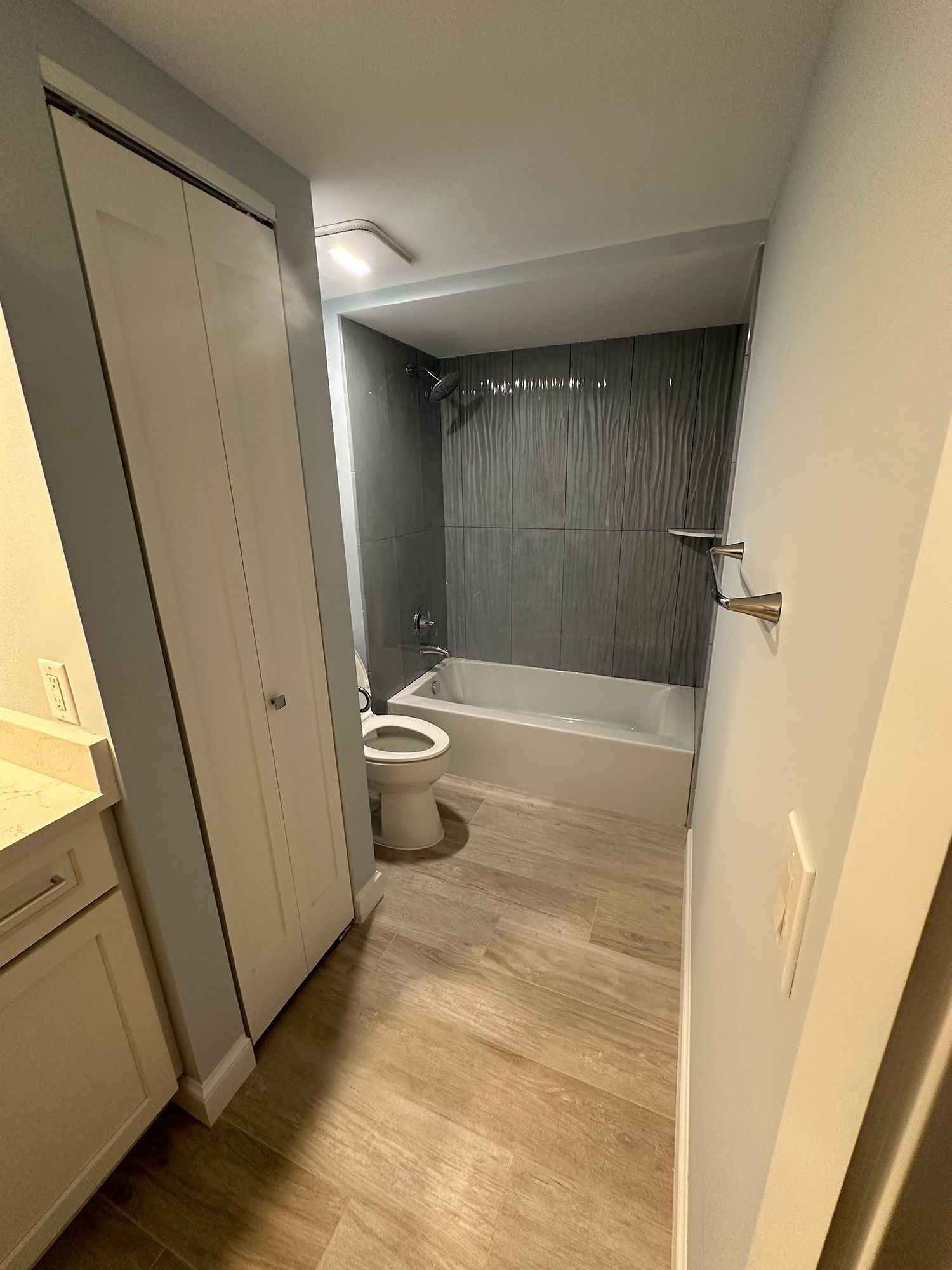 Bathroom with wood-look flooring, white tub, toilet, and vanity. Gray tile shower surround and light blue walls.