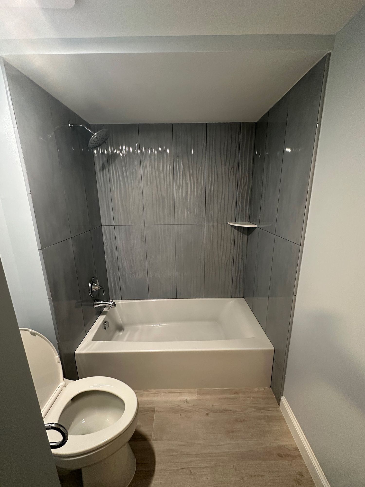 Bathroom with a white tub, toilet, and gray tiled walls. Light wood-look flooring.
