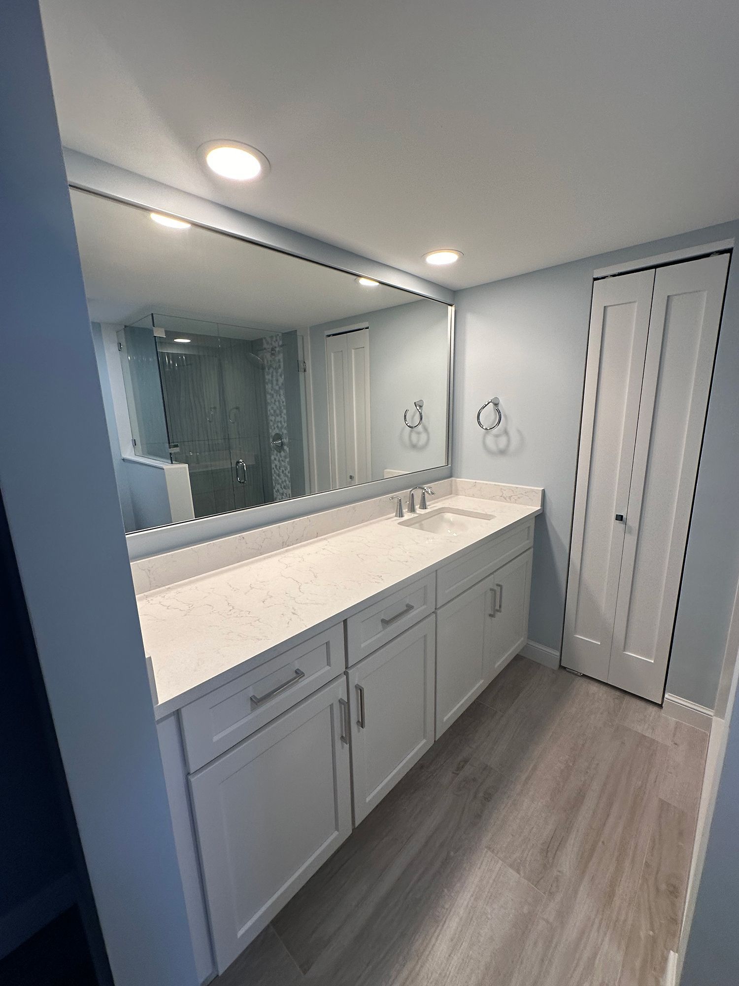 Bathroom with large mirror over white vanity, light blue walls, and light wood-look floor.