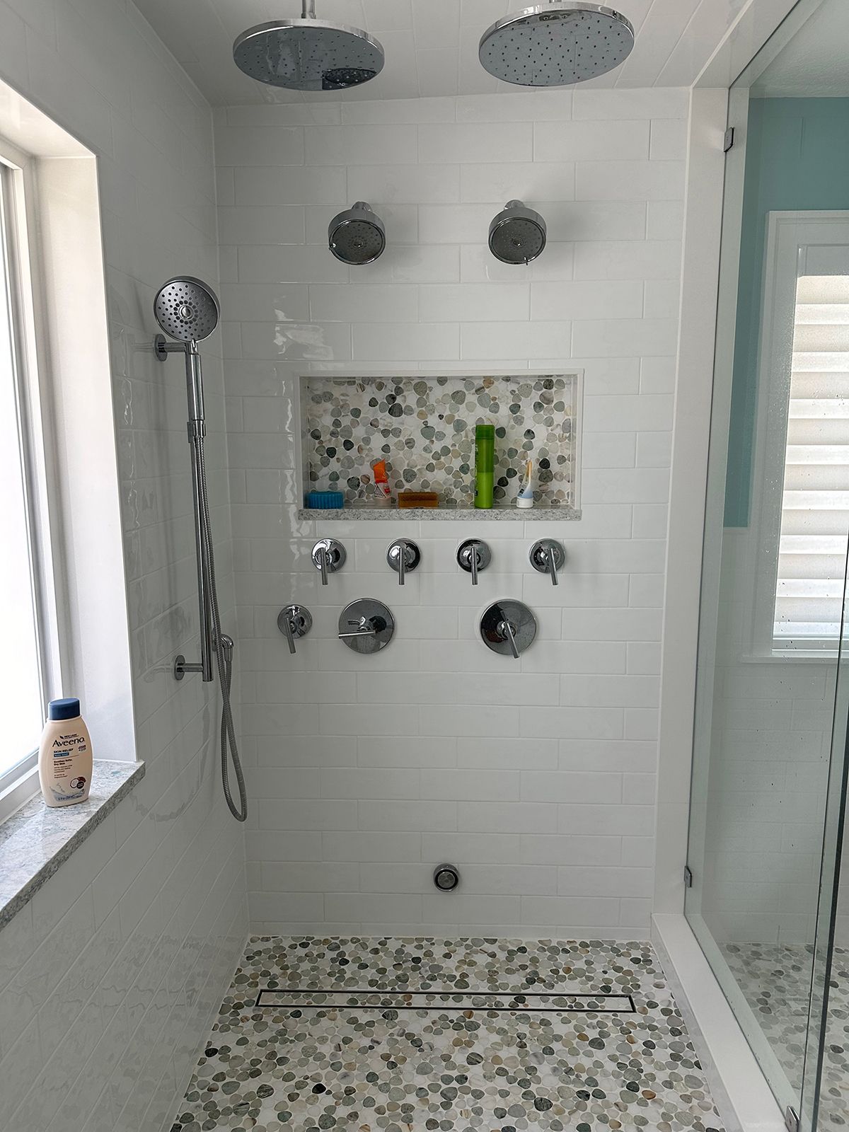 White tiled shower with multiple showerheads, a recessed shelf, and a glass door.