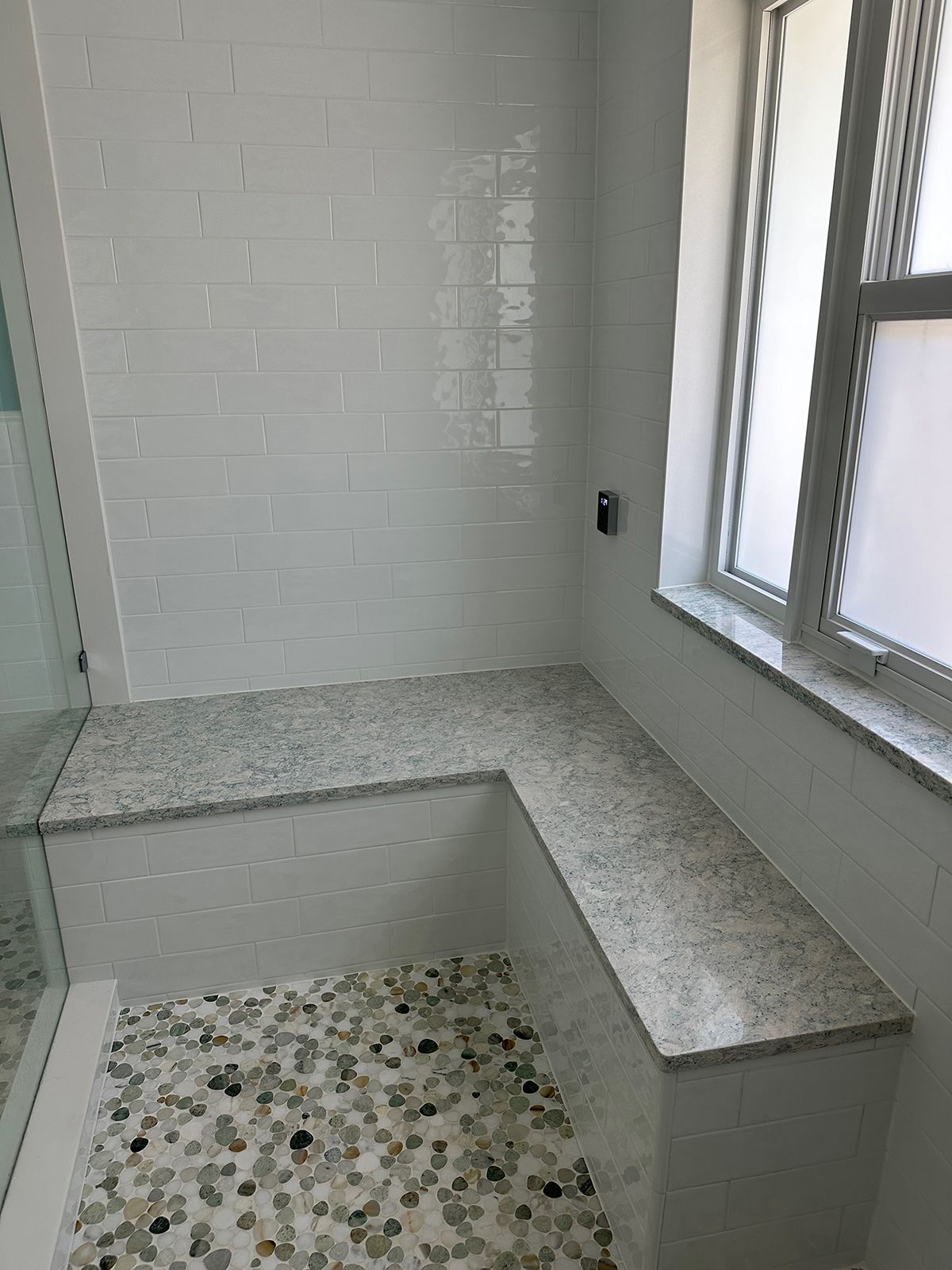 White tiled shower with granite bench and pebble floor, next to a window.