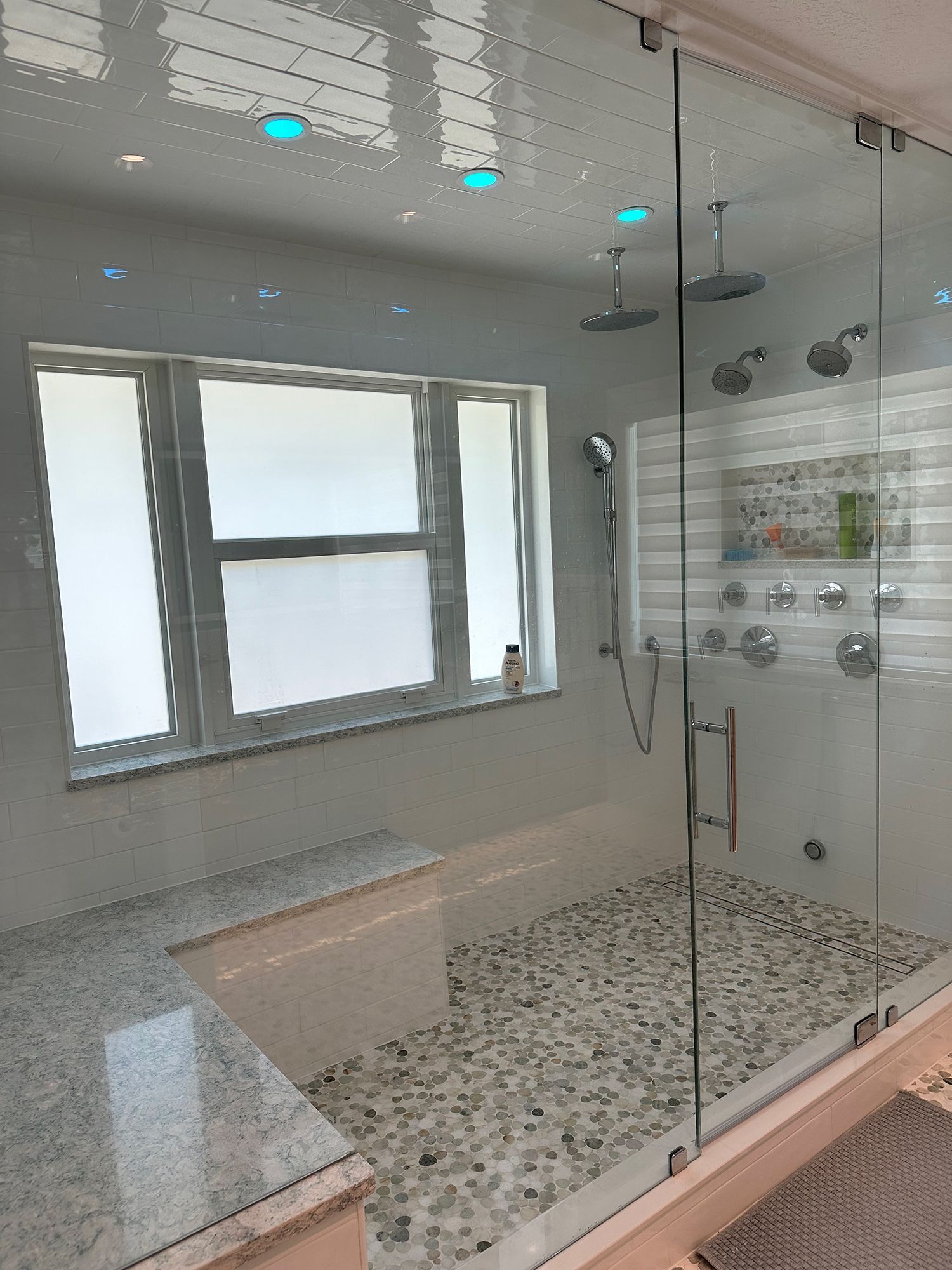 Modern shower with glass doors, pebble floor, built-in bench, window, and multiple shower heads.