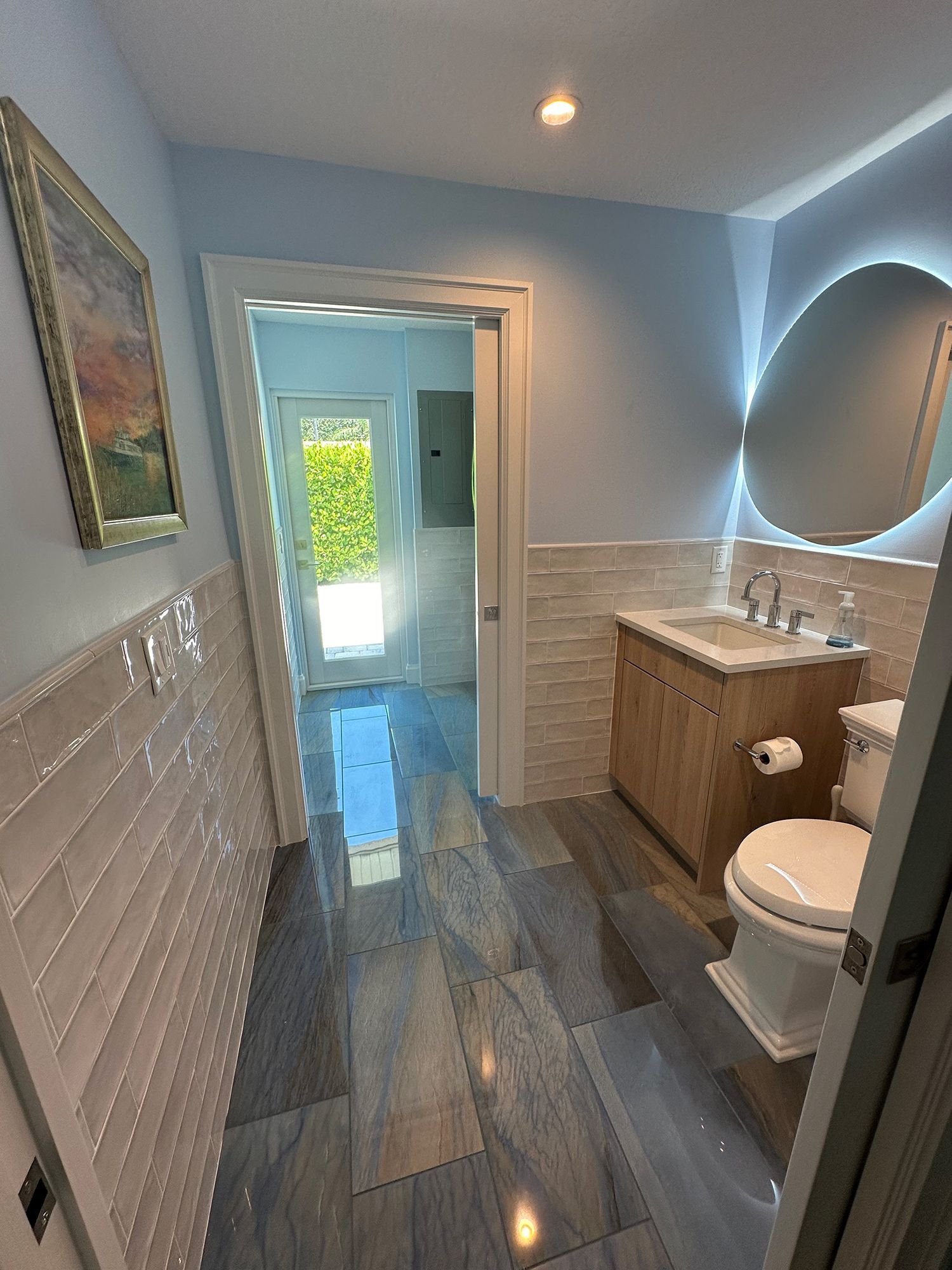 Bathroom with blue walls, wood vanity, white toilet, patterned floor, and doorway to outside.