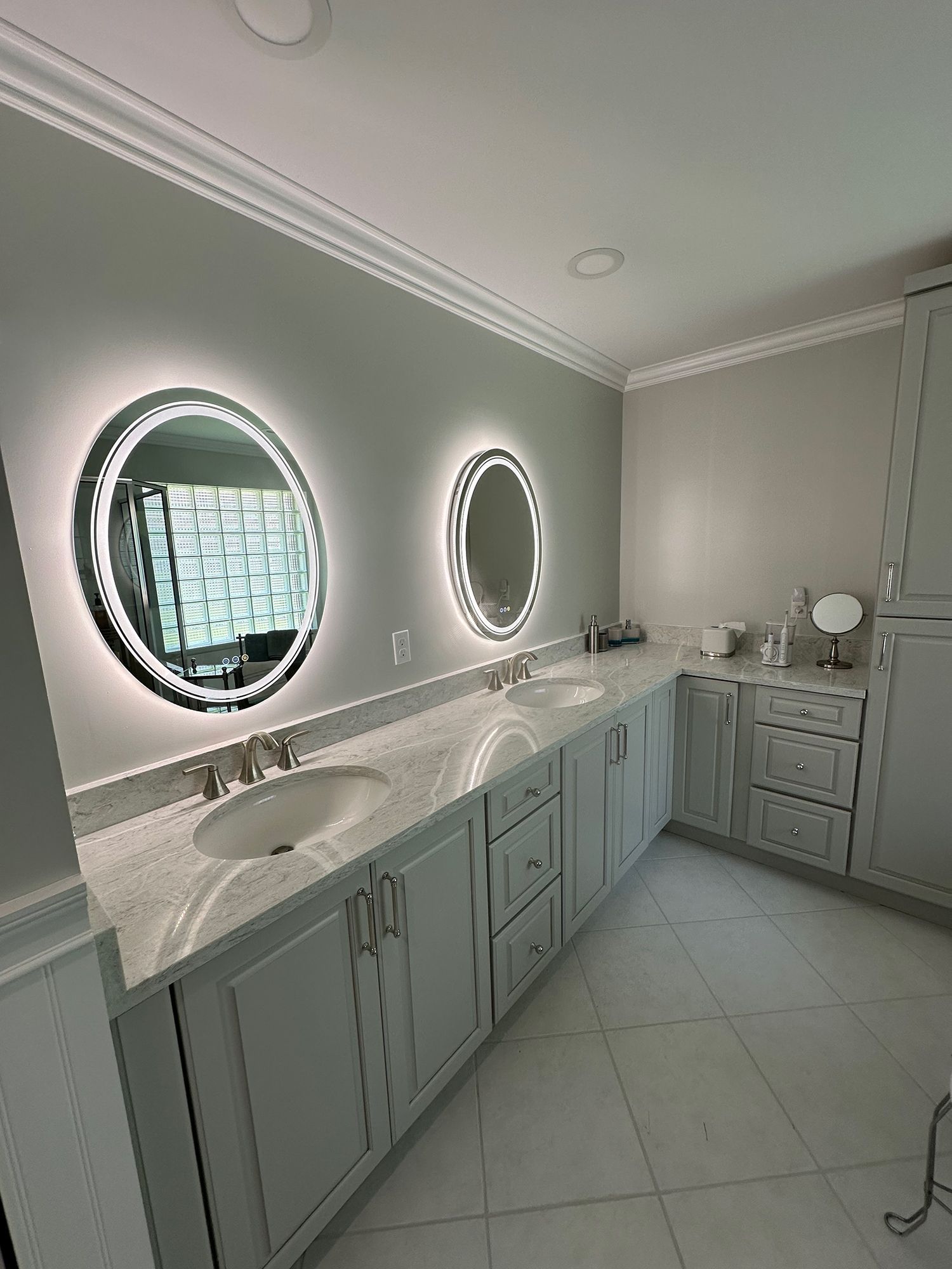 Bathroom with two illuminated round mirrors above sinks and light gray cabinets.