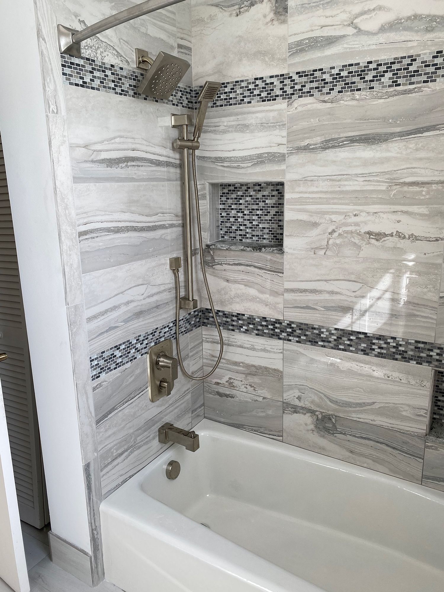 Bathroom with tiled walls, showerhead, and bathtub. Features grey and blue tiles.