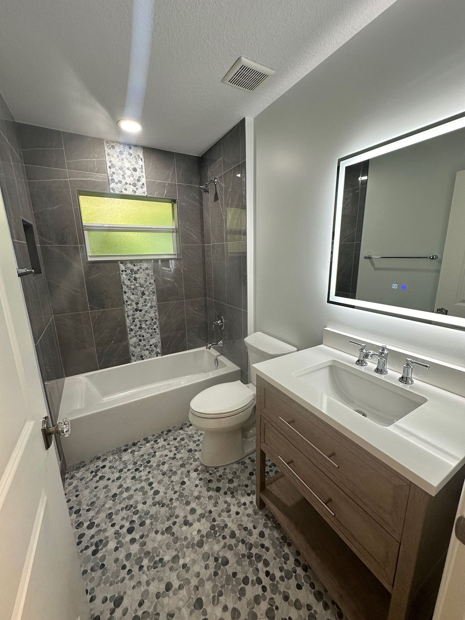Bathroom with gray and pebble-tiled floor, bathtub, toilet, vanity with lighted mirror, and shower.