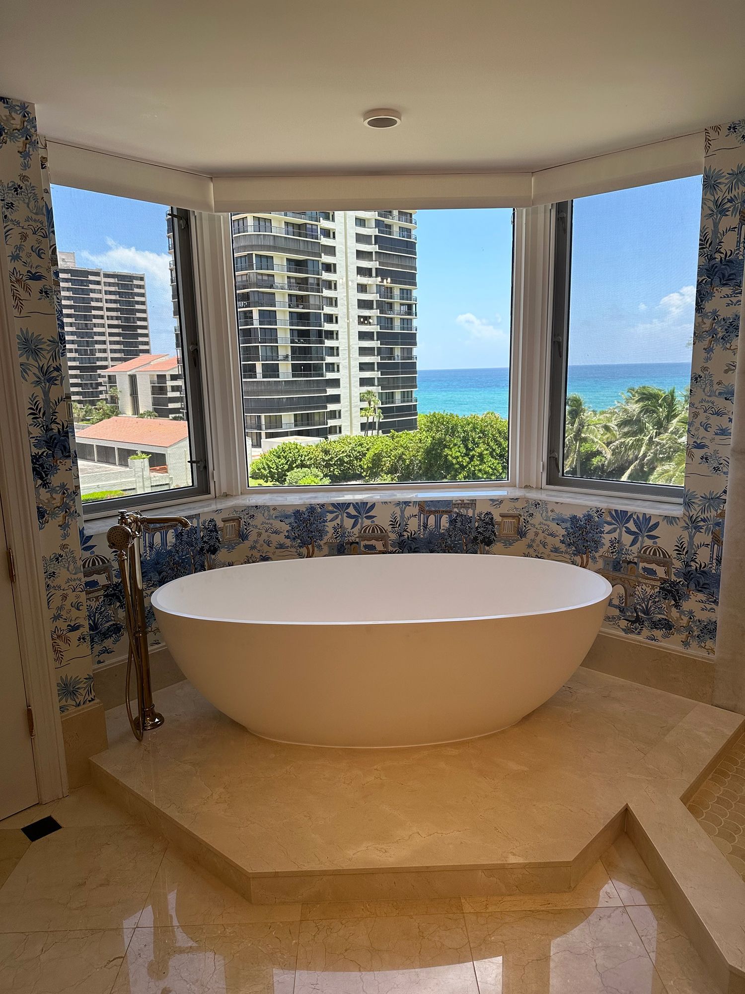 Bathroom with a white oval bathtub in front of a bay window with ocean and city views. Blue floral wallpaper.